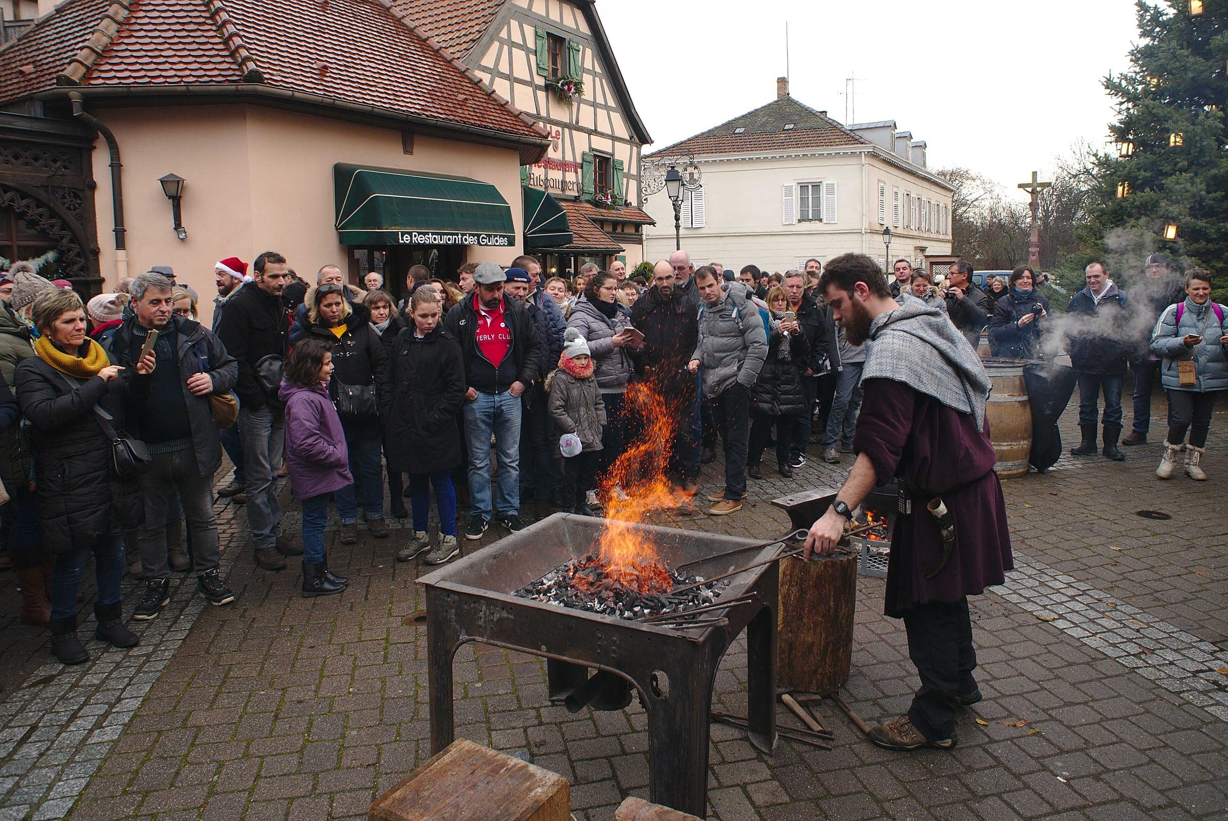 A blacksmith in medieval costume works a glowing forge for a crowd at the Ribeauvillé Christmas market.