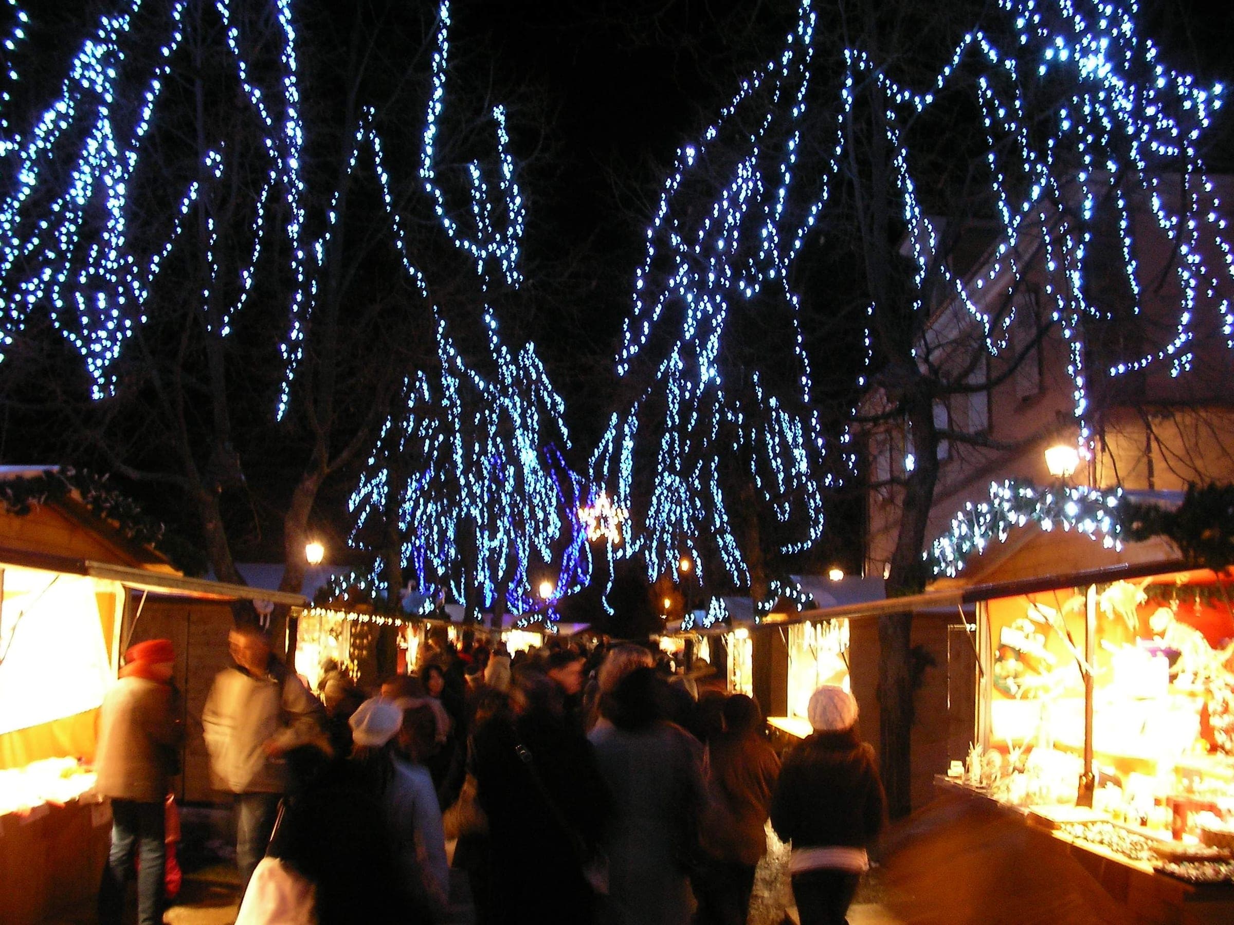 Blue lights hang from the trees over a lane of lit market chalets in Riquewihr at night.