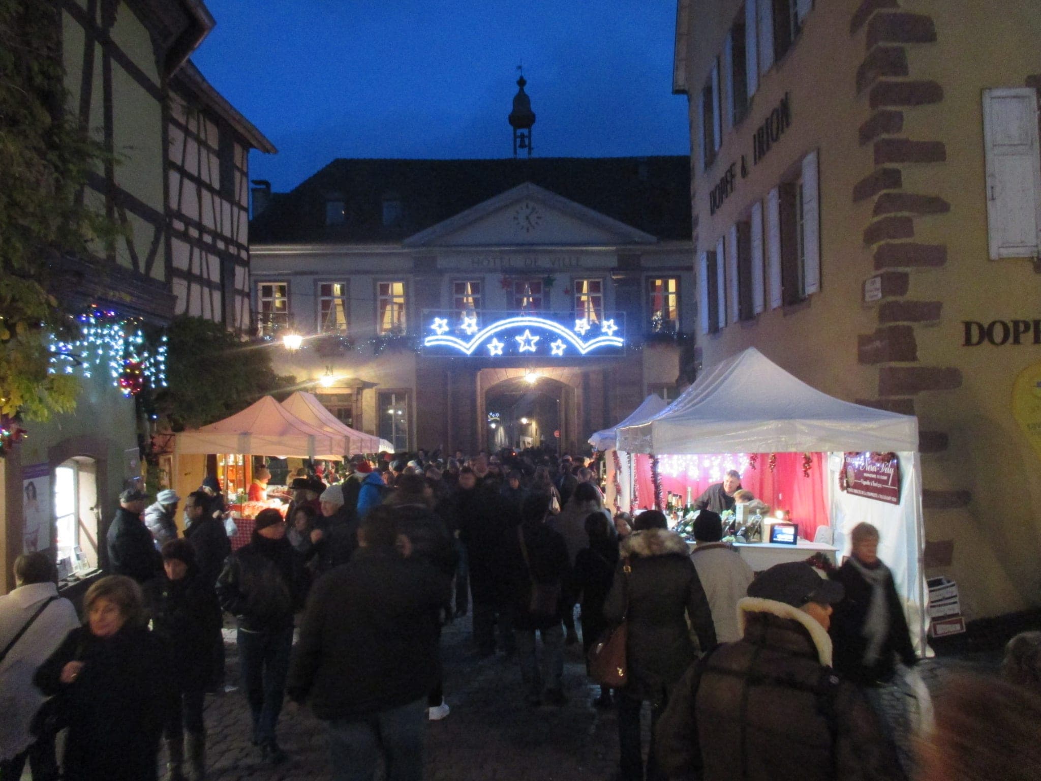 Visitors fill the main street of Riquewihr at dusk as lights glow on the town hall at the end of the market.
