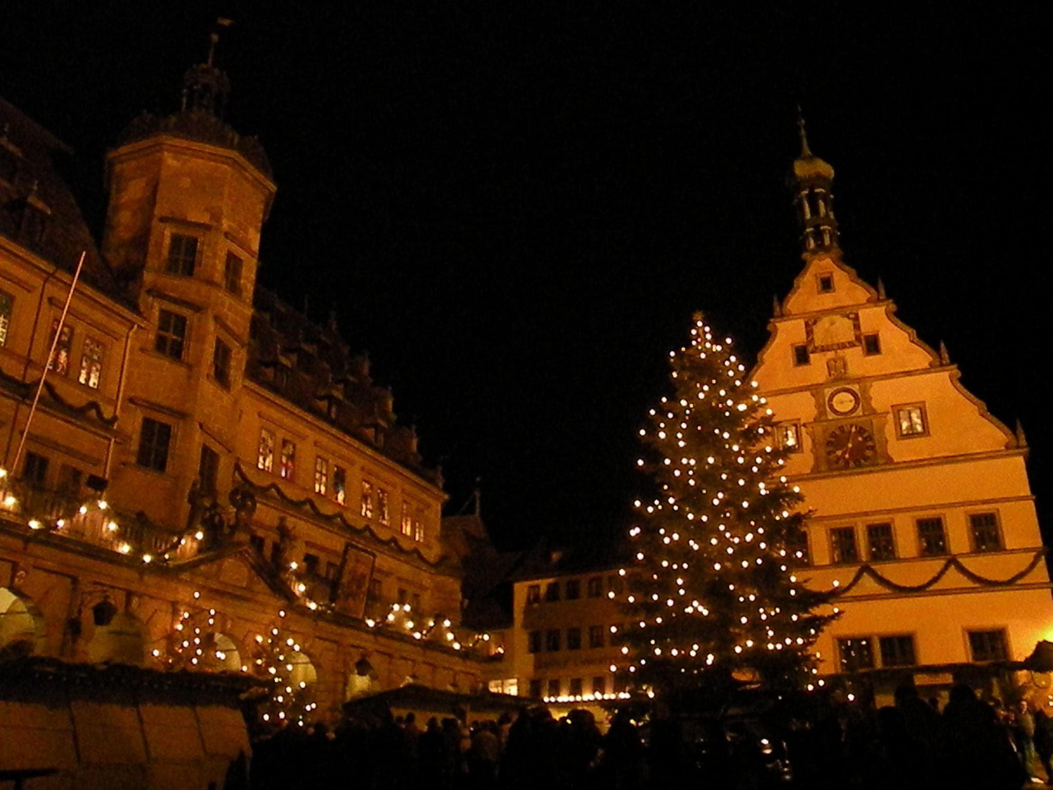 A tall lit tree stands between the Rathaus and the Ratstrinkstube during Rothenburg's Reiterlesmarkt at night