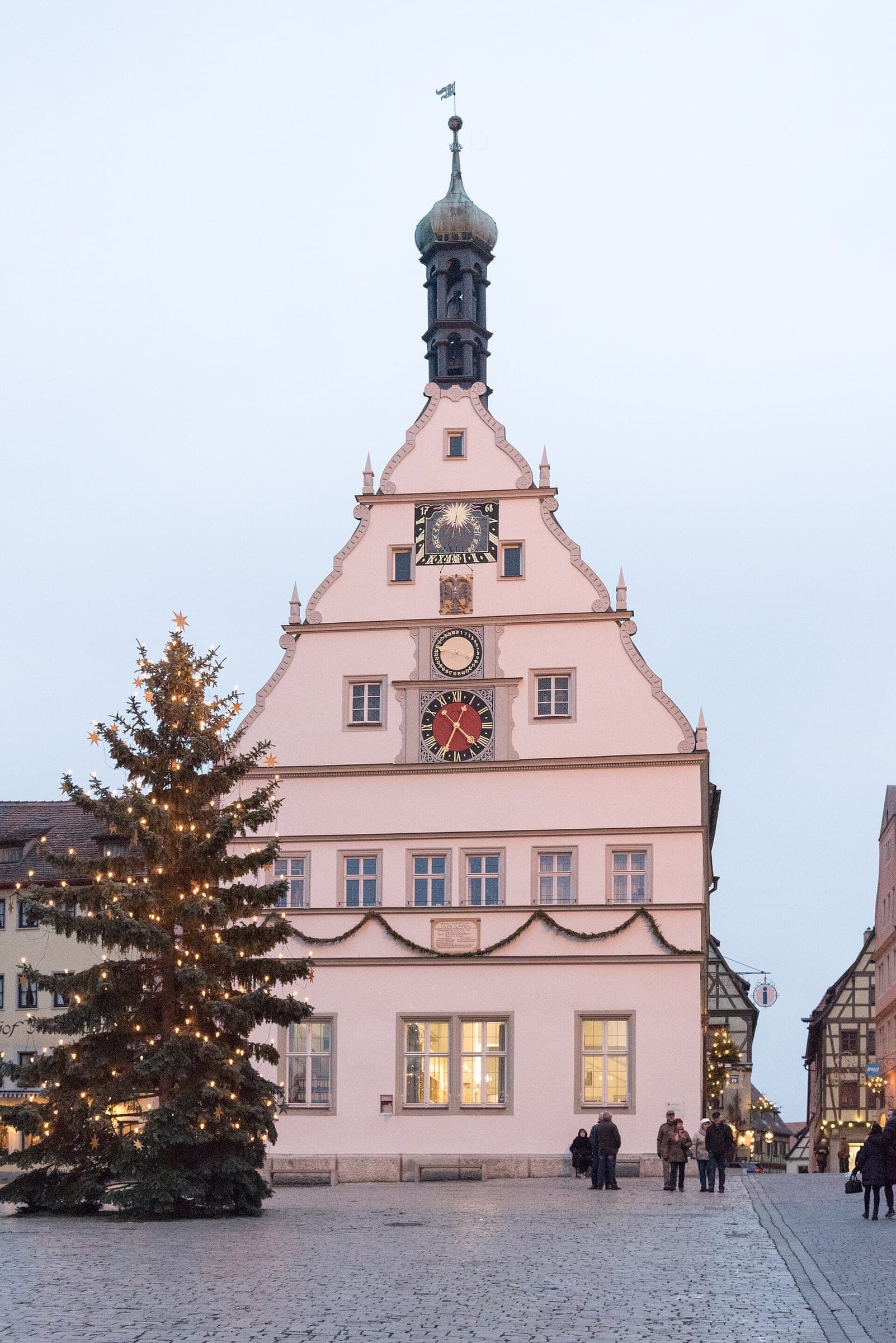A lit Christmas tree stands before the clock gable of the Ratstrinkstube on Rothenburg's Marktplatz at dusk