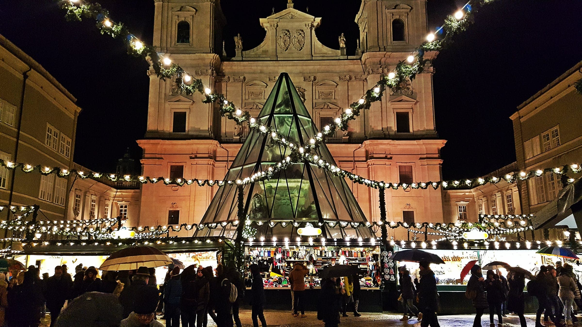 Lit stalls of the Salzburg Christkindlmarkt fill the Domplatz at night below the cathedral facade