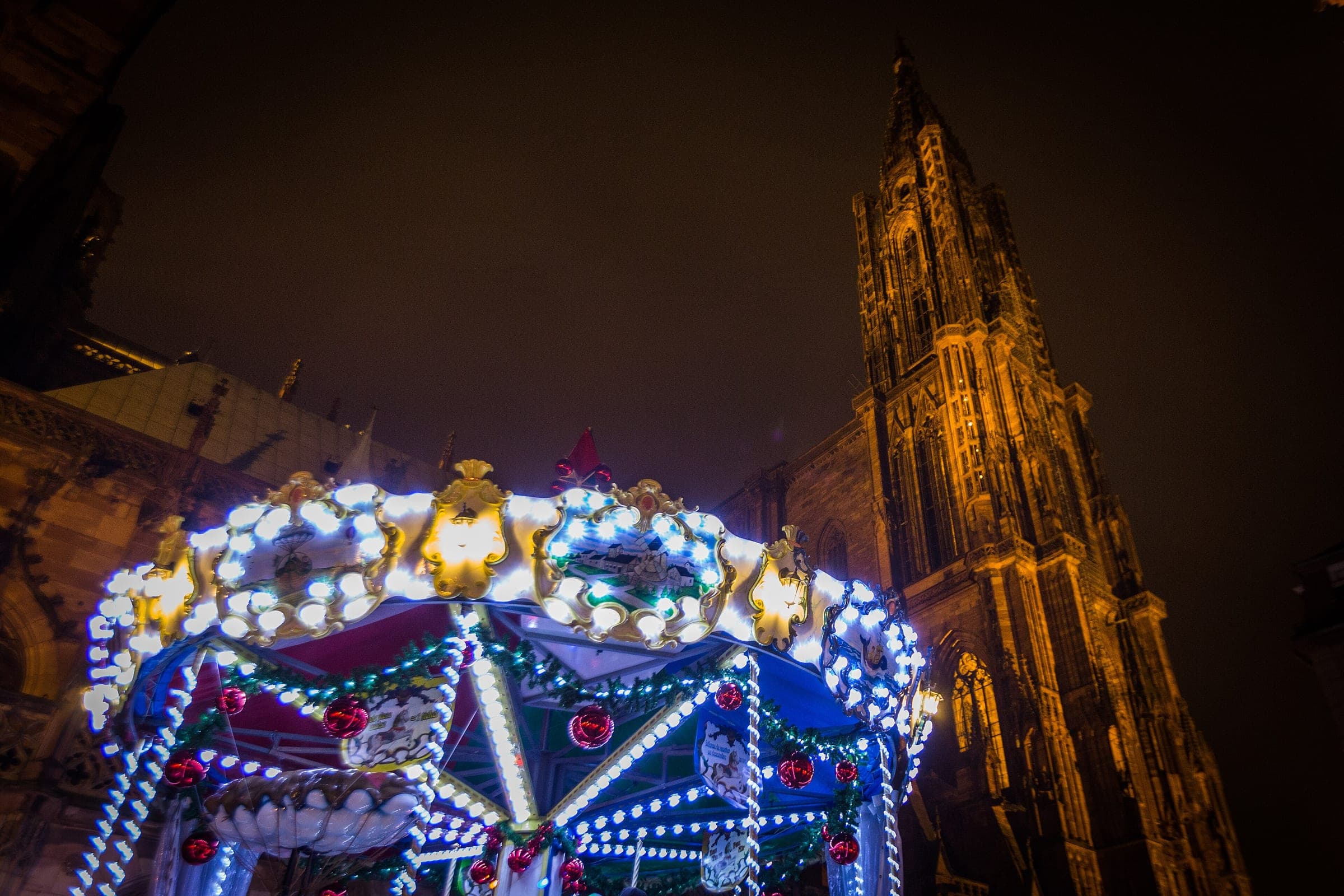 A lit carousel turns on Place de la Cathédrale in Strasbourg below the floodlit spire of the cathedral.