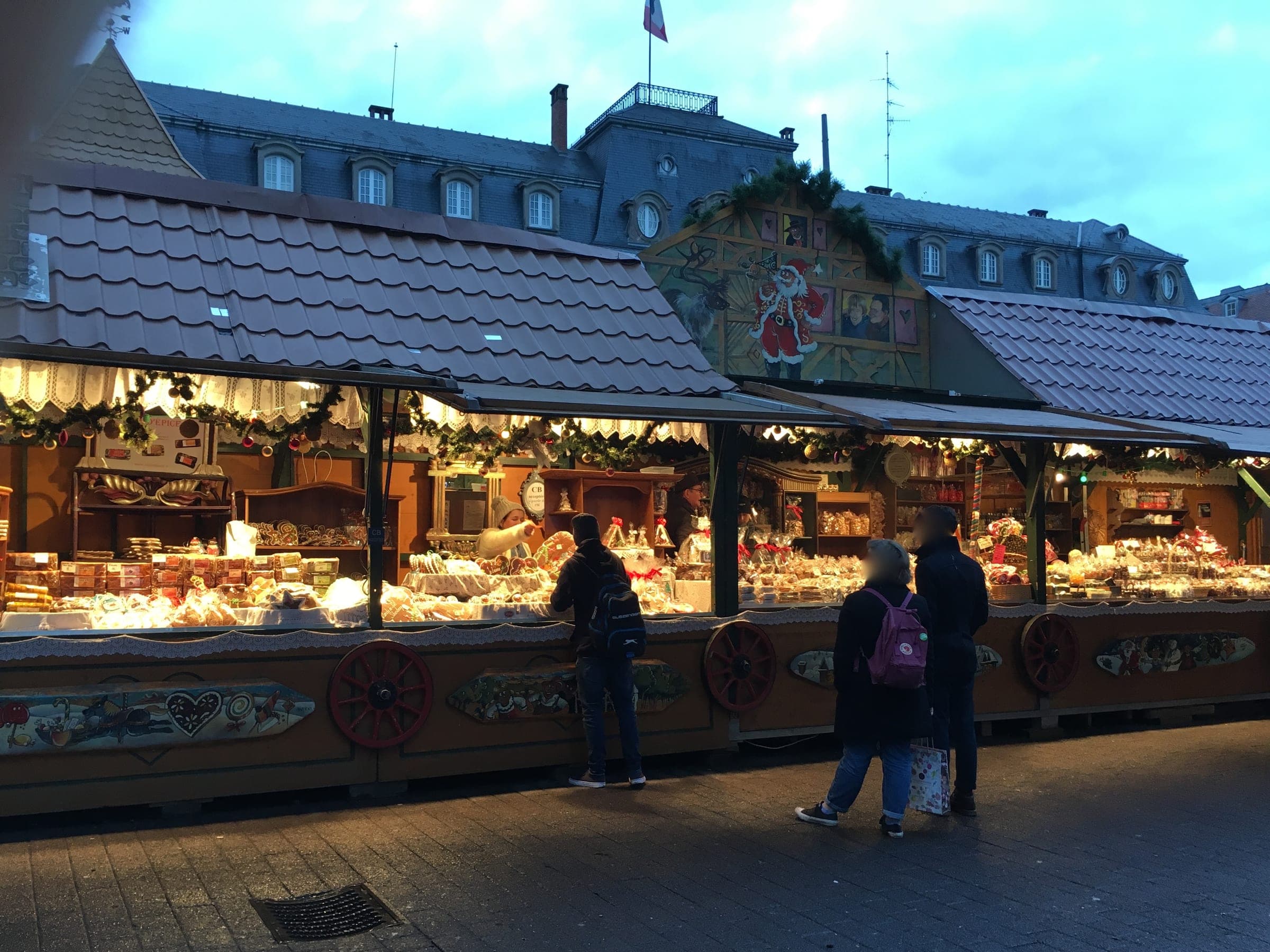 Shoppers stop at a long lit stall on Place Broglie in Strasbourg as the evening sky turns blue.