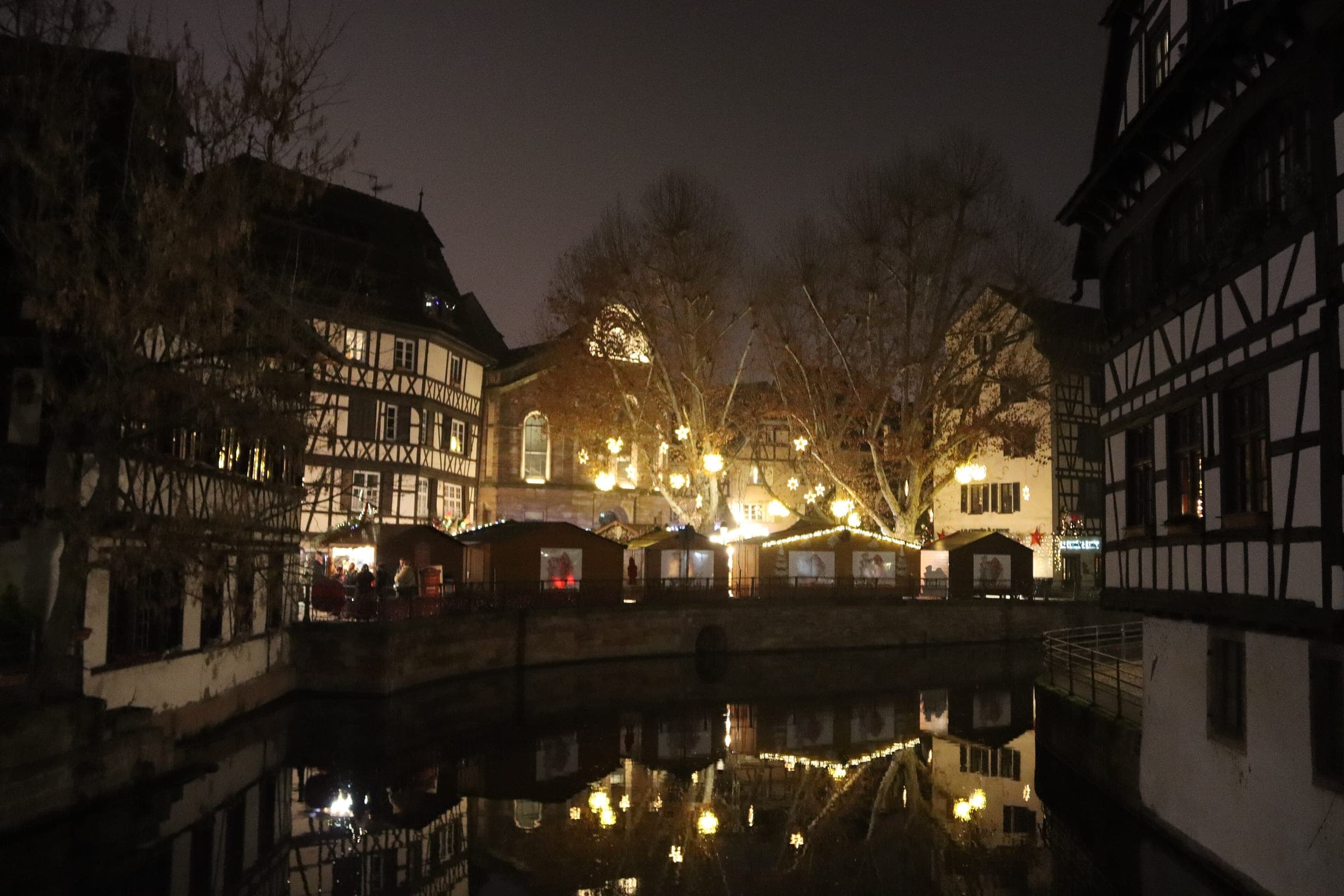 Market chalets glow along the canal in Strasbourg's Petite France as the lights reflect in the water at night.