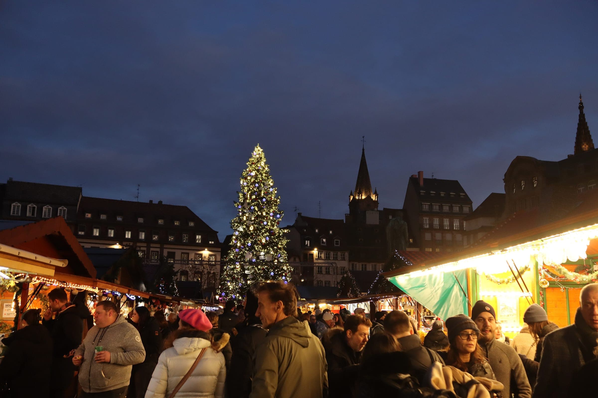 Crowds fill the lit stalls on Place Kléber in Strasbourg while the Grand Sapin glows against a blue evening sky.