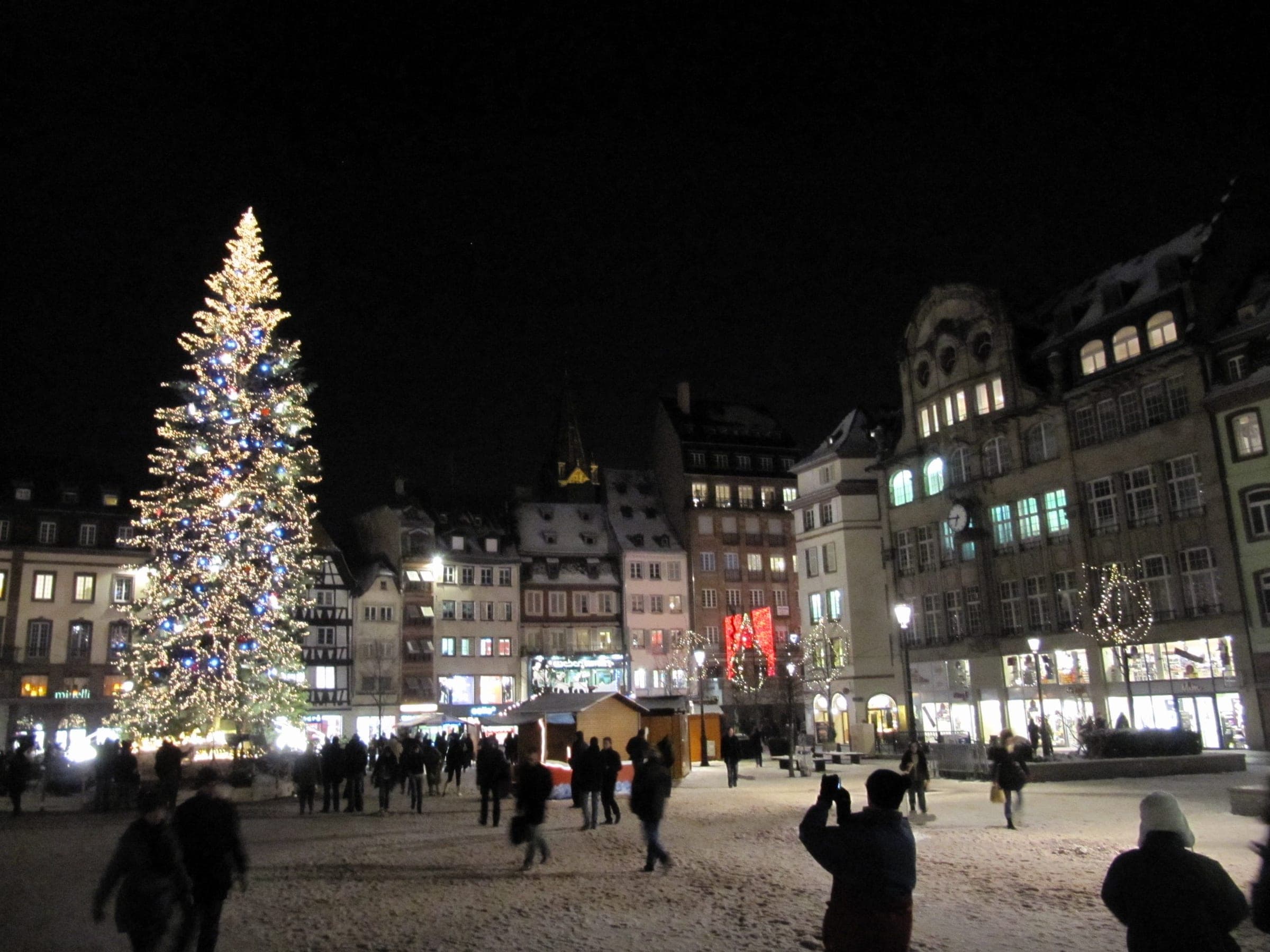 Snow lies on Place Kléber in Strasbourg as people cross the square toward the lit Grand Sapin at night.