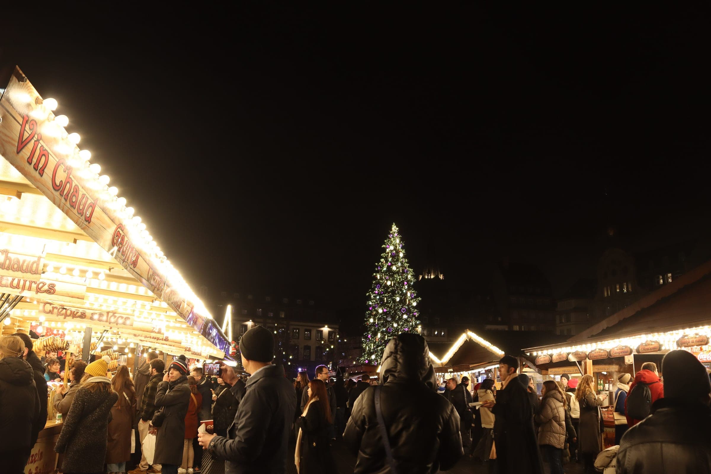 People crowd the vin chaud stalls on Place Kléber in Strasbourg with the Grand Sapin lit behind them.