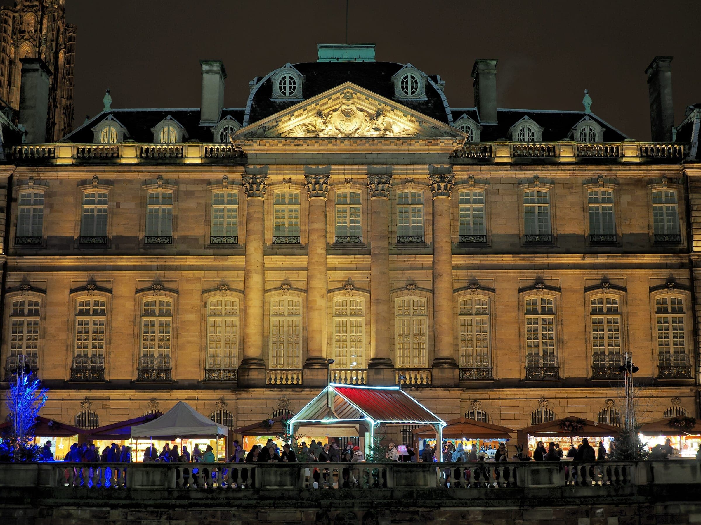Market stalls glow on the Terrasse Rohan below the floodlit river facade of the Palais Rohan in Strasbourg.