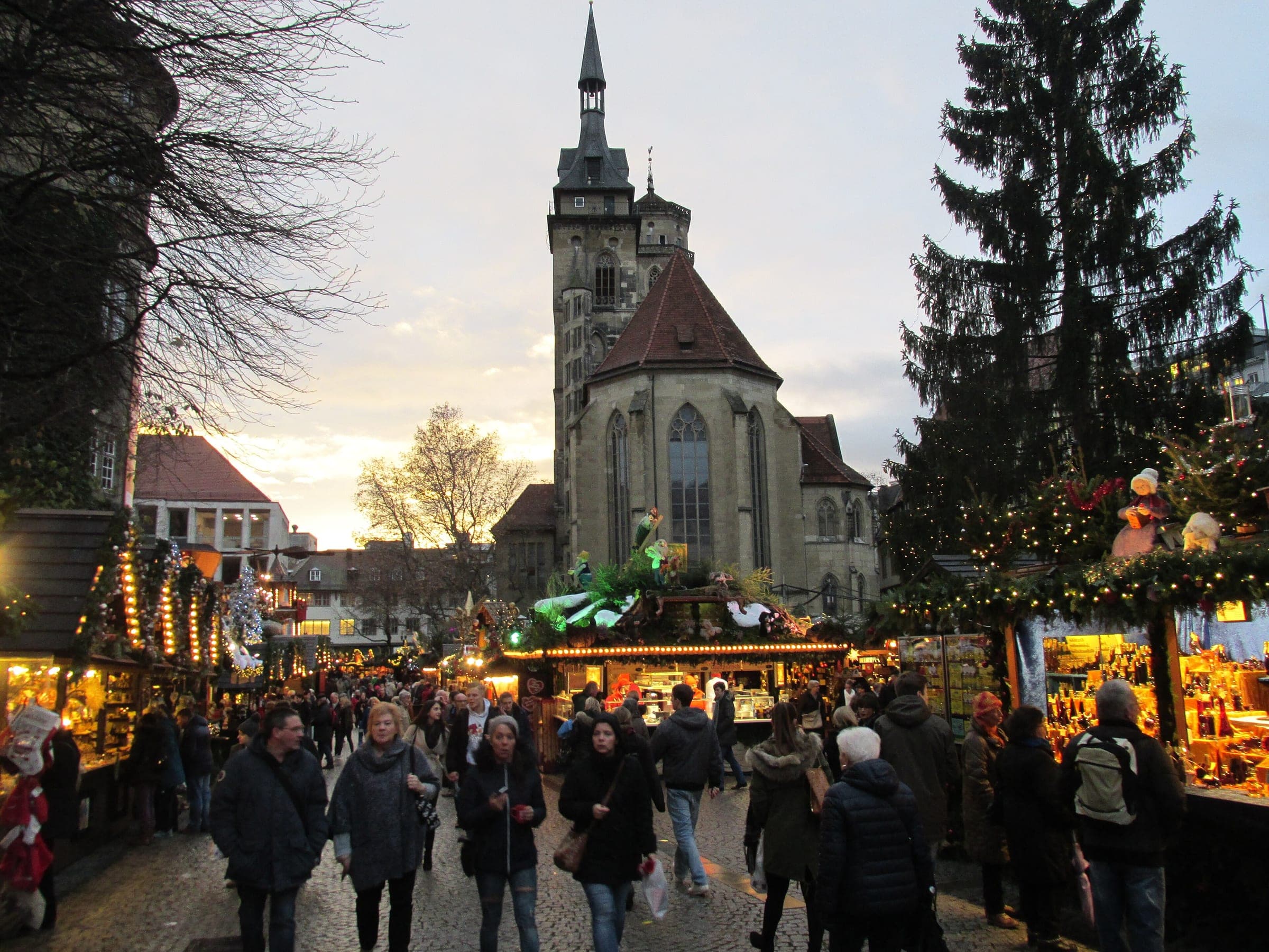 Shoppers walk between market stalls toward the Stiftskirche in Stuttgart as the December sky fades.