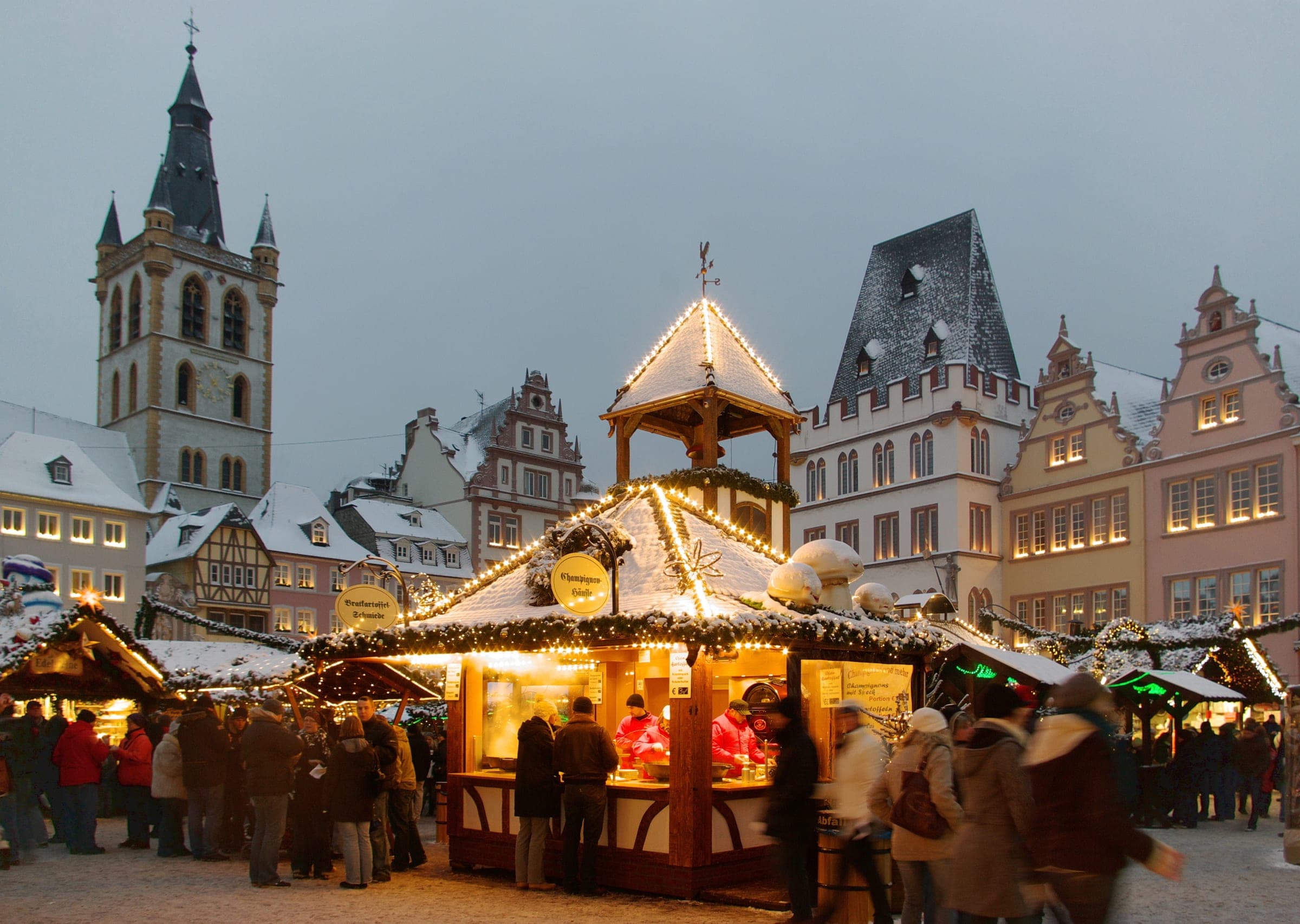 Snow lies on the lit stalls of the Trier Christmas market below St Gangolf on the Hauptmarkt at dusk