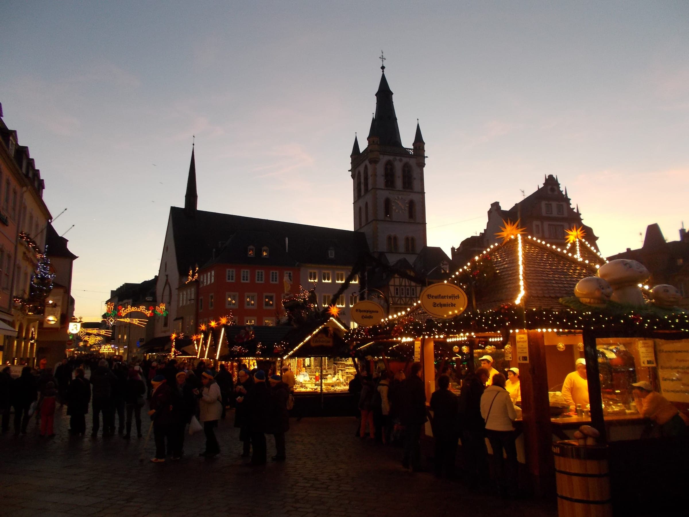 The tower of St Gangolf catches the last light above market crowds on Trier's Hauptmarkt