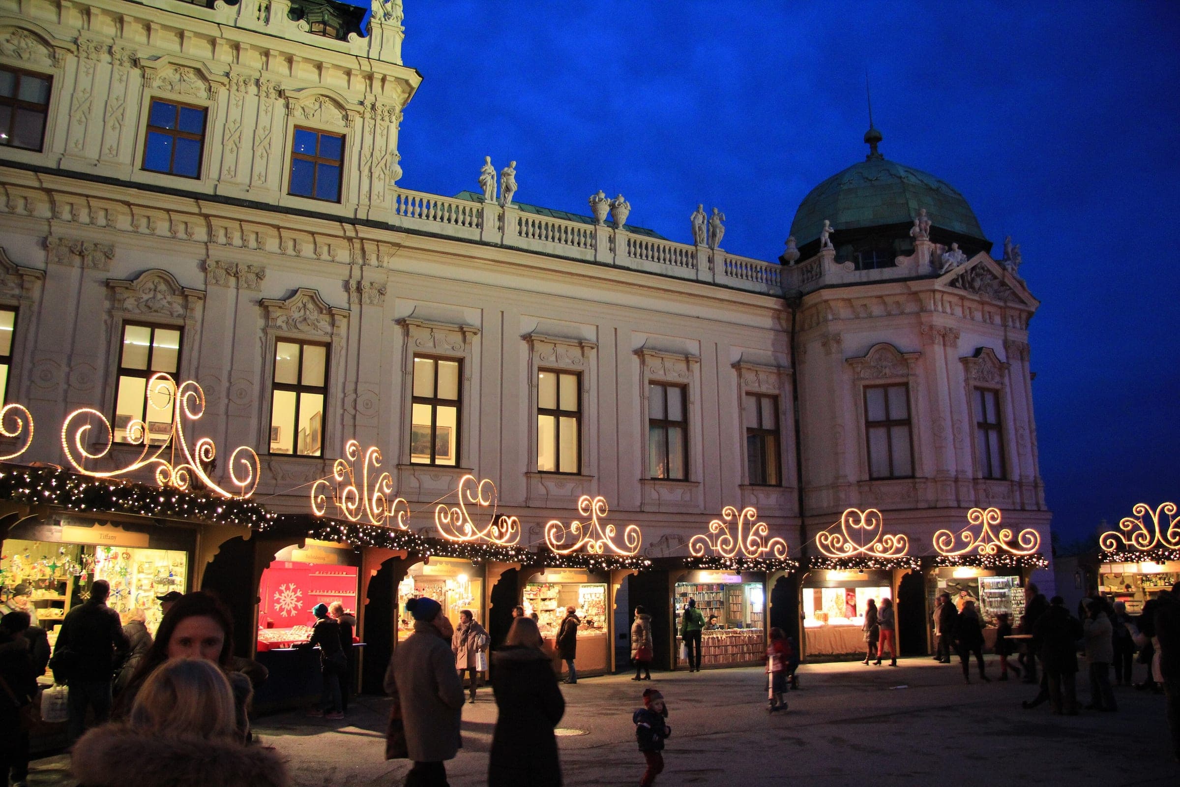 Visitors stroll past the scrolled light arches of the stalls below the Upper Belvedere in Vienna at dusk.