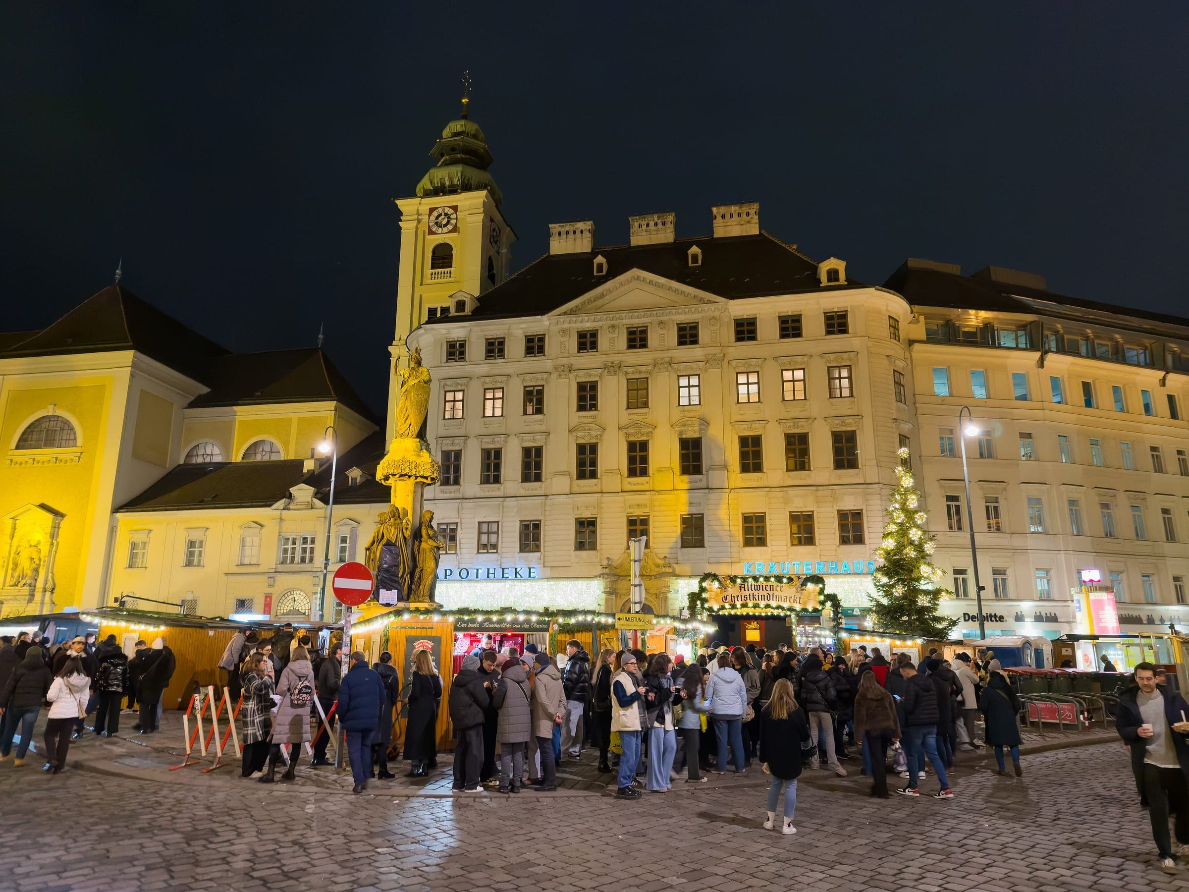 Crowds fill the Freyung market in Vienna at night below the lit tree and the Schottenkirche tower.