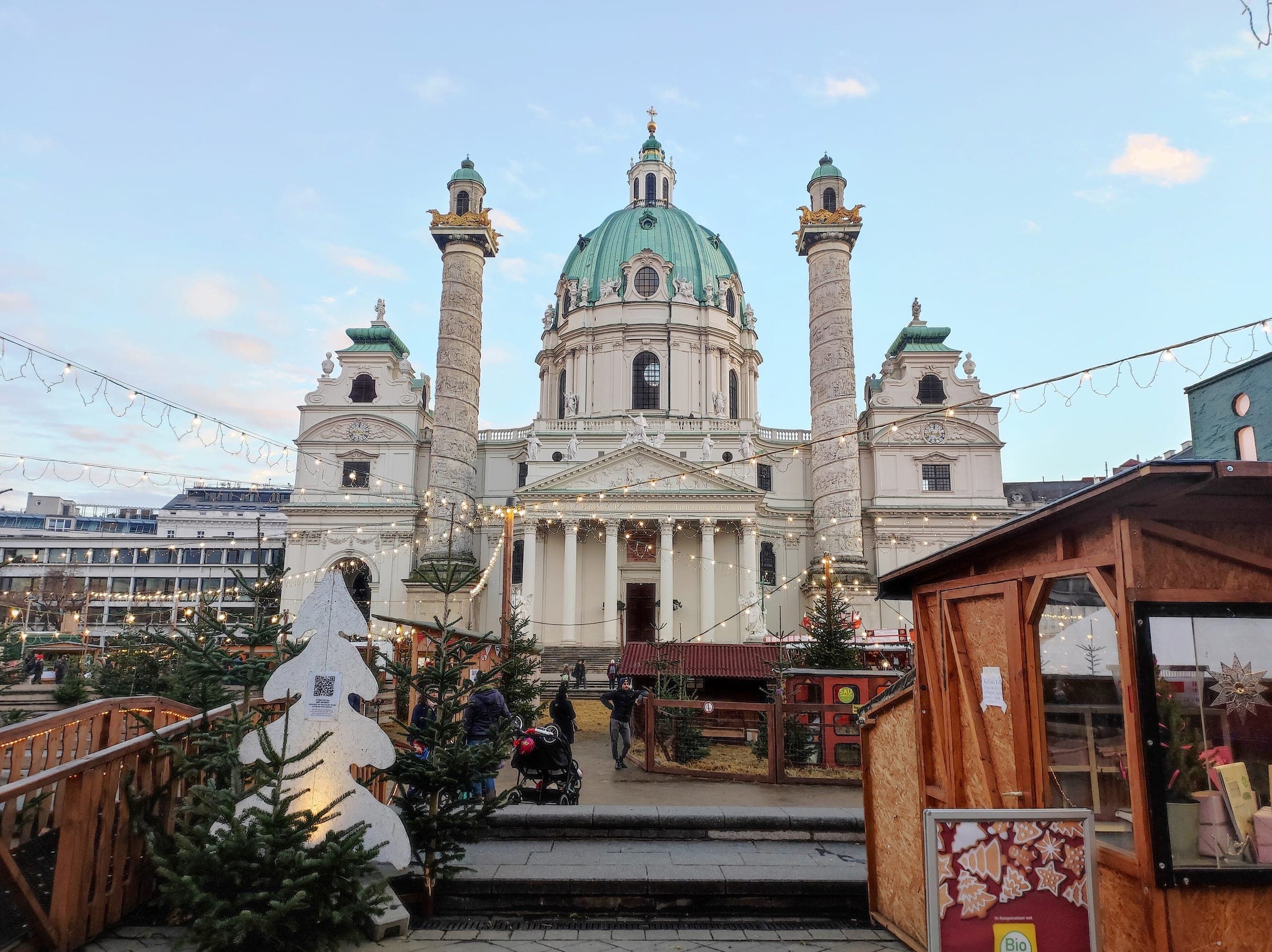 The Karlskirche rises behind a snowy stall roof at the Karlsplatz Christmas market in Vienna by day.