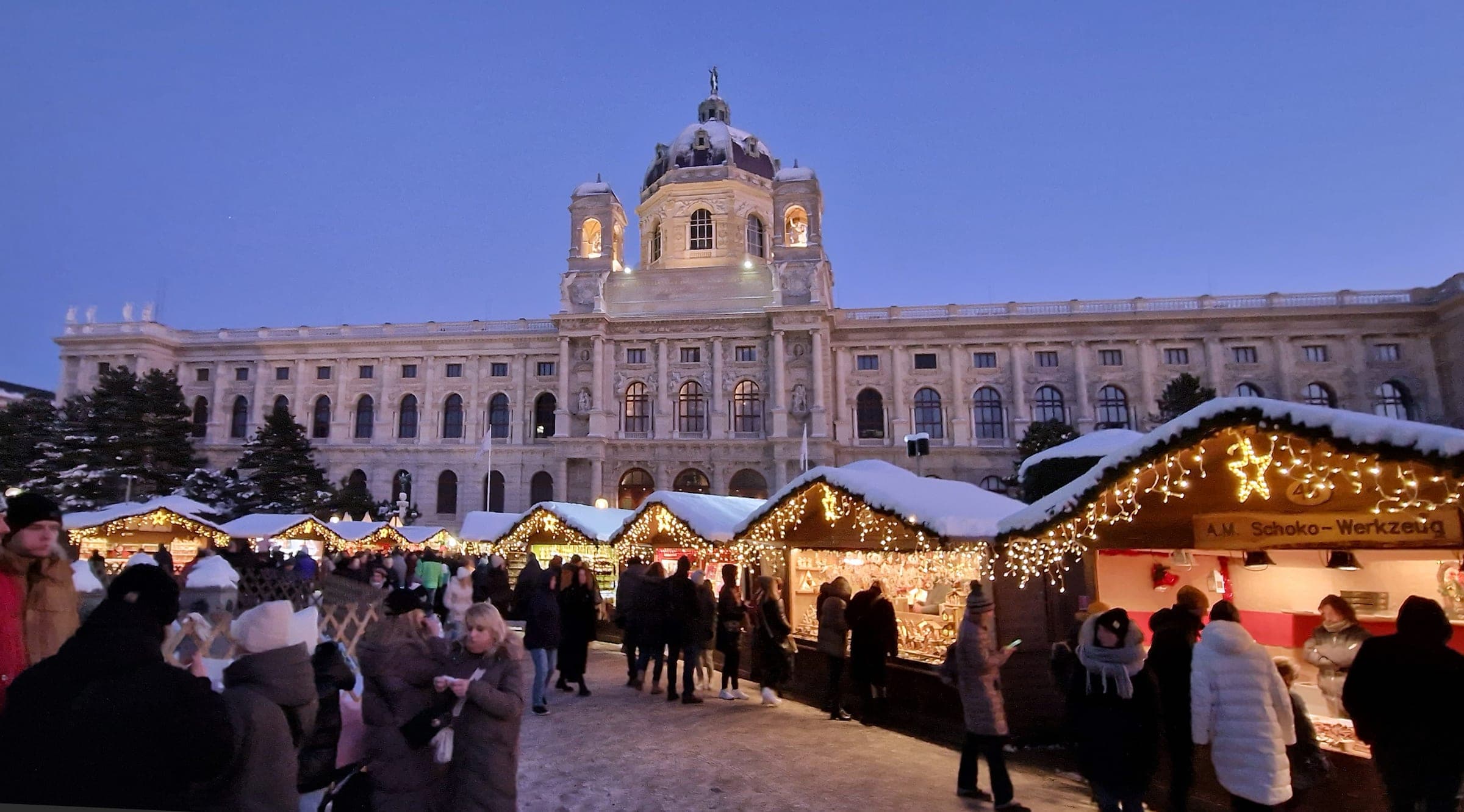 Lit stalls with snowy roofs fill Maria-Theresien-Platz in Vienna below the Kunsthistorisches Museum at dusk.