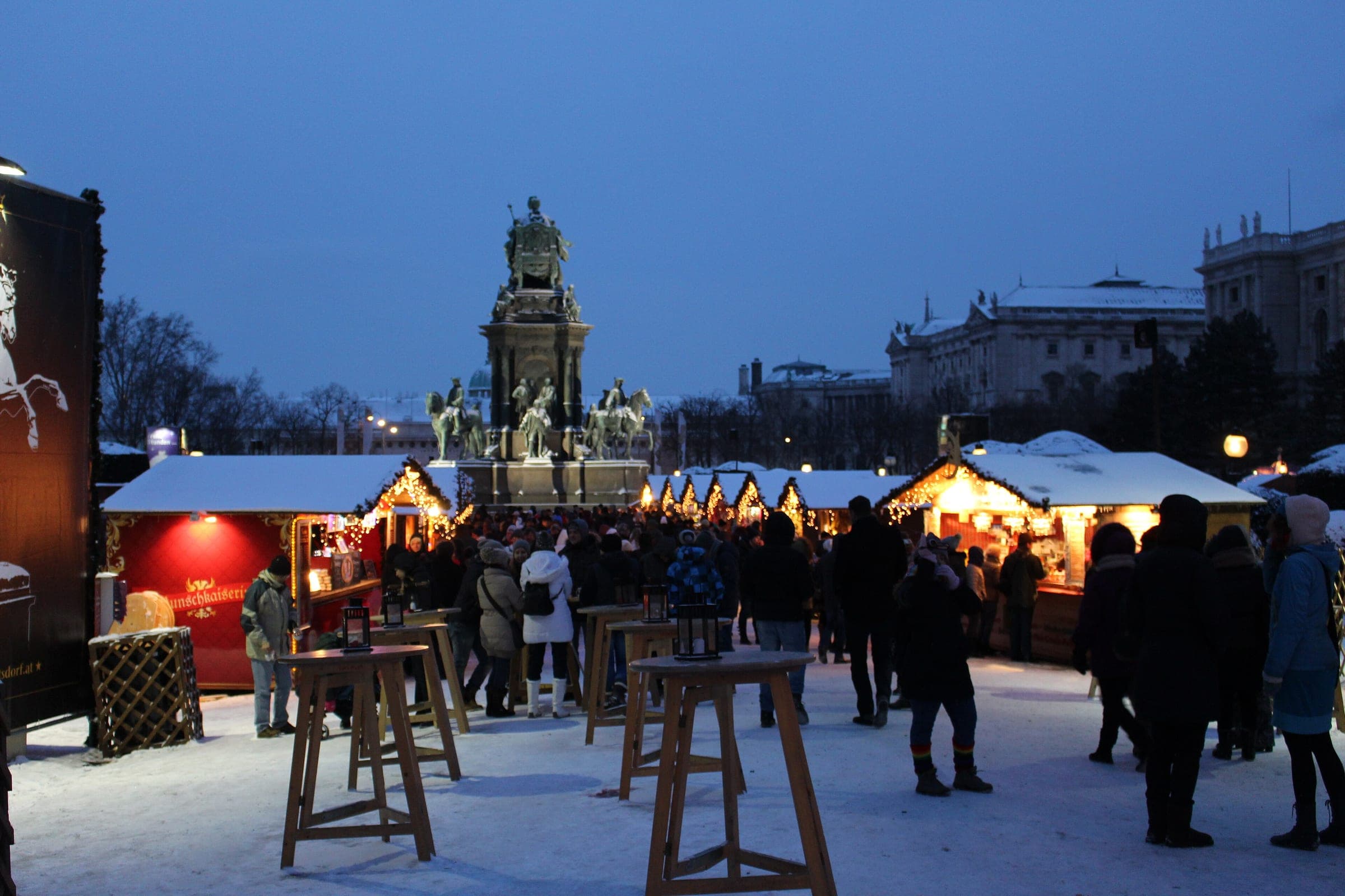 Snow covers the Maria-Theresien-Platz Christmas village in Vienna at dusk below the Maria Theresa monument.