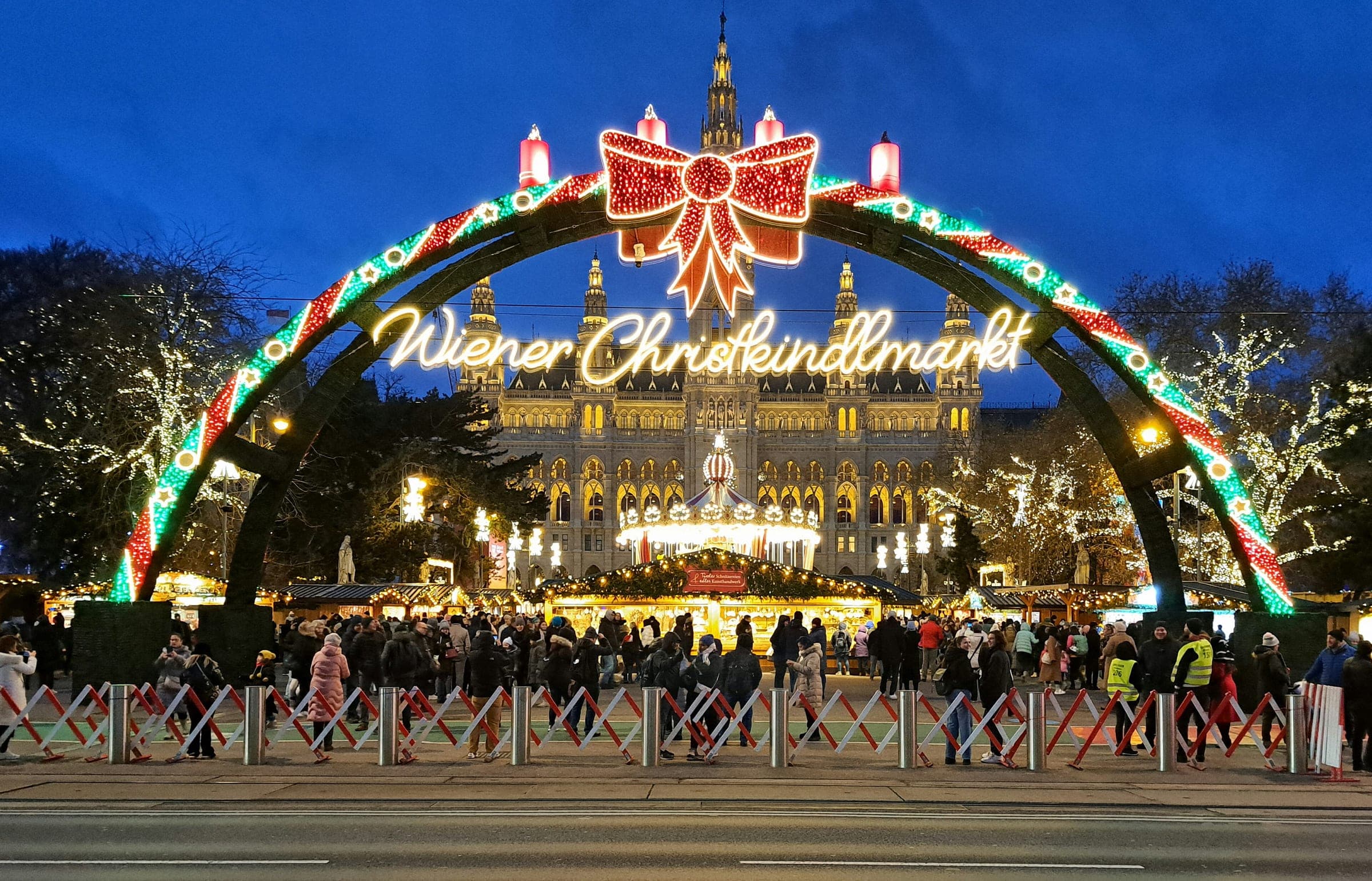 The lit entrance arch of the Wiener Christkindlmarkt frames Vienna City Hall at dusk on the Rathausplatz.