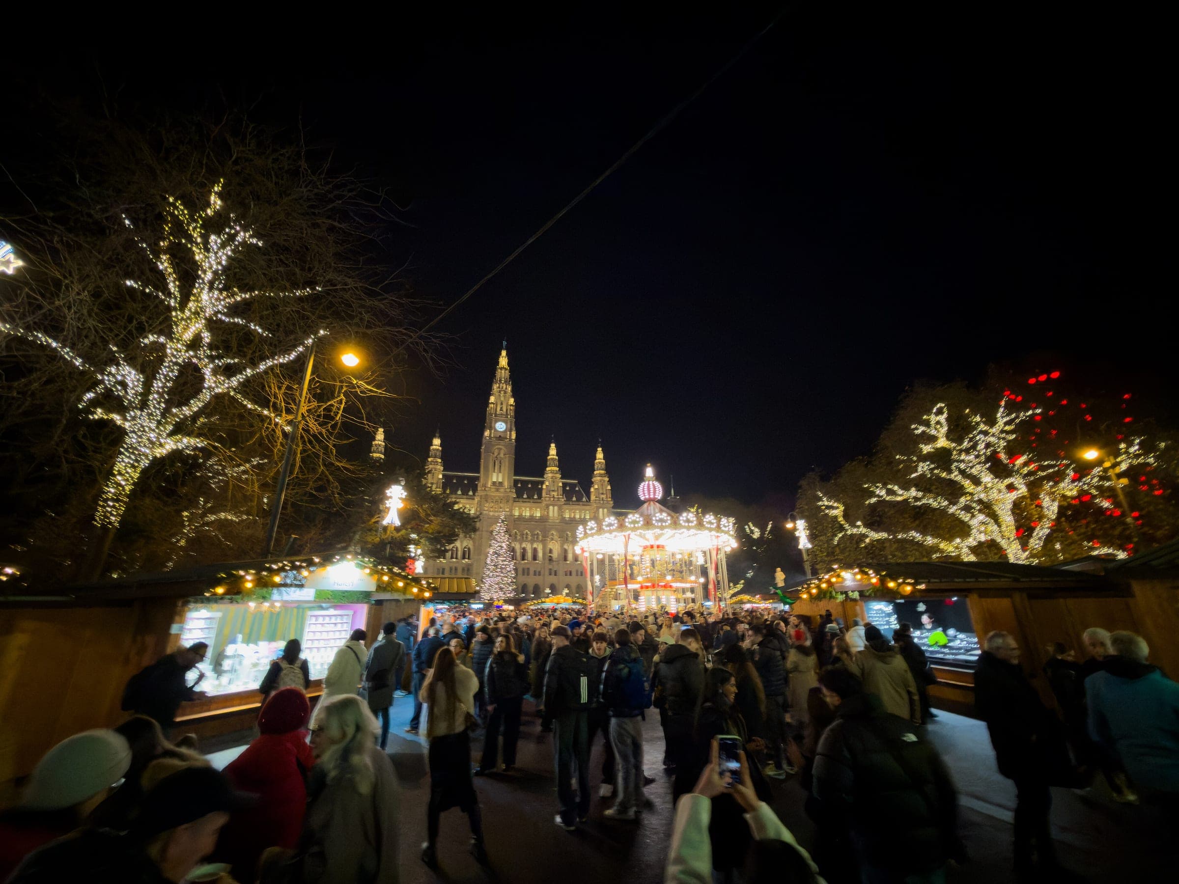 Crowds walk between lit trees toward the carousel and City Hall at the Rathausplatz market in Vienna.