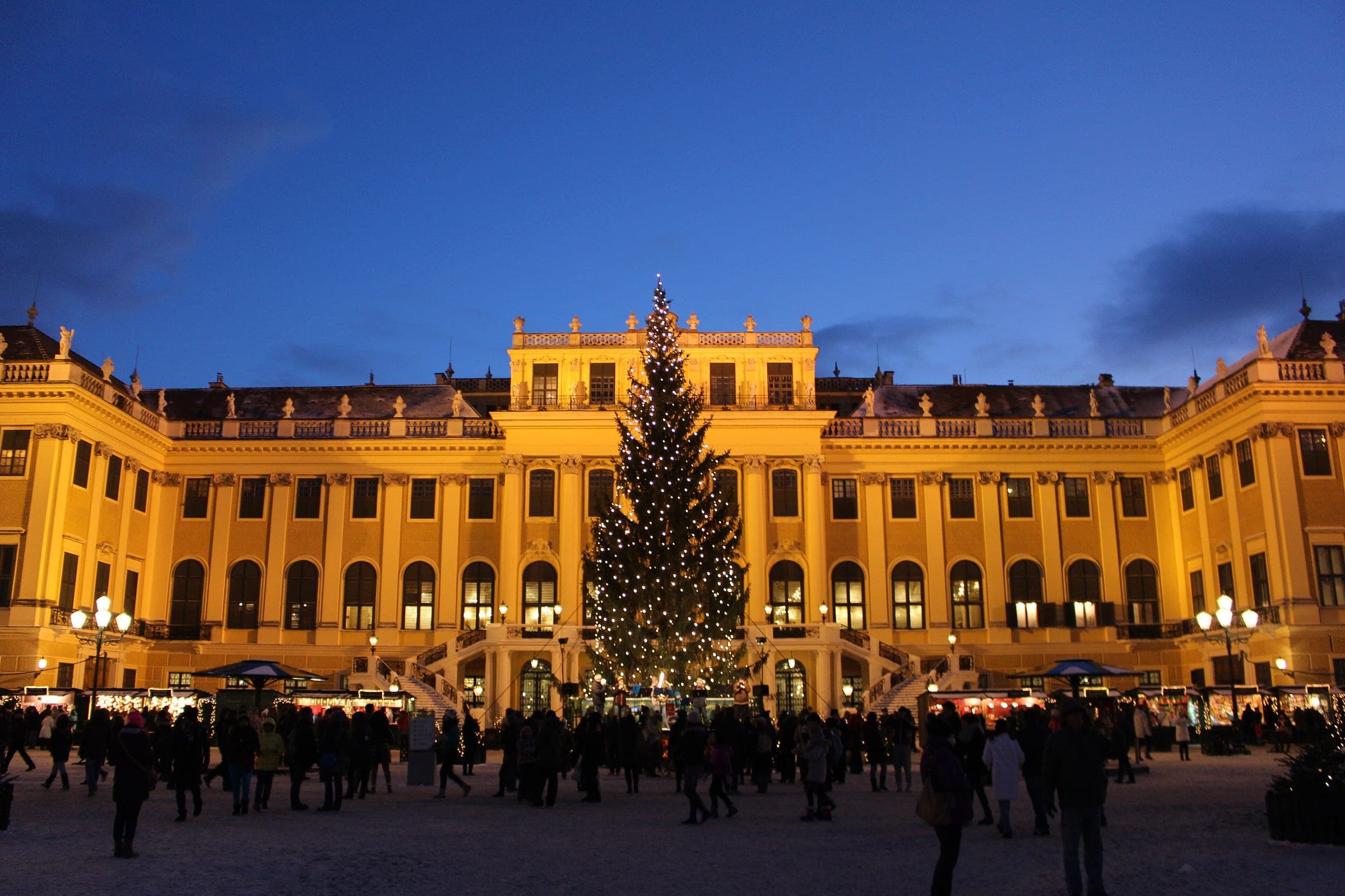 A lit Christmas tree stands before Schonbrunn Palace in Vienna as the market fills the forecourt at blue hour.
