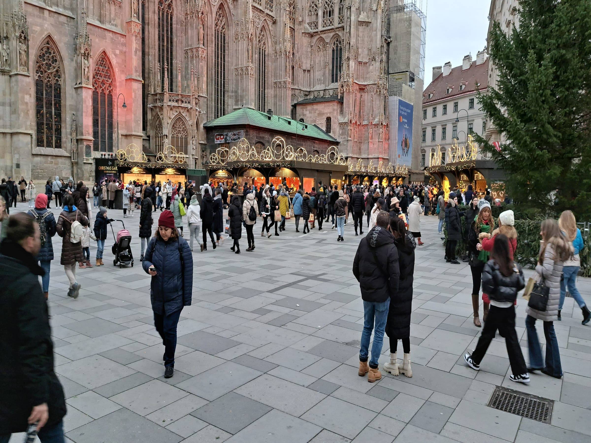 Lit stalls with gold scroll crowns line Stephansplatz in Vienna at dusk beside St. Stephen's Cathedral.