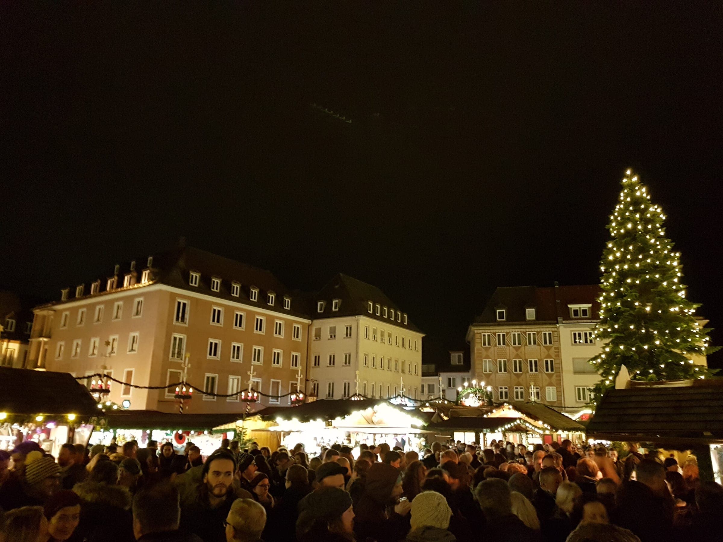 A crowd fills the Würzburg Christmas market below a lit tree and the facades of the Marktplatz