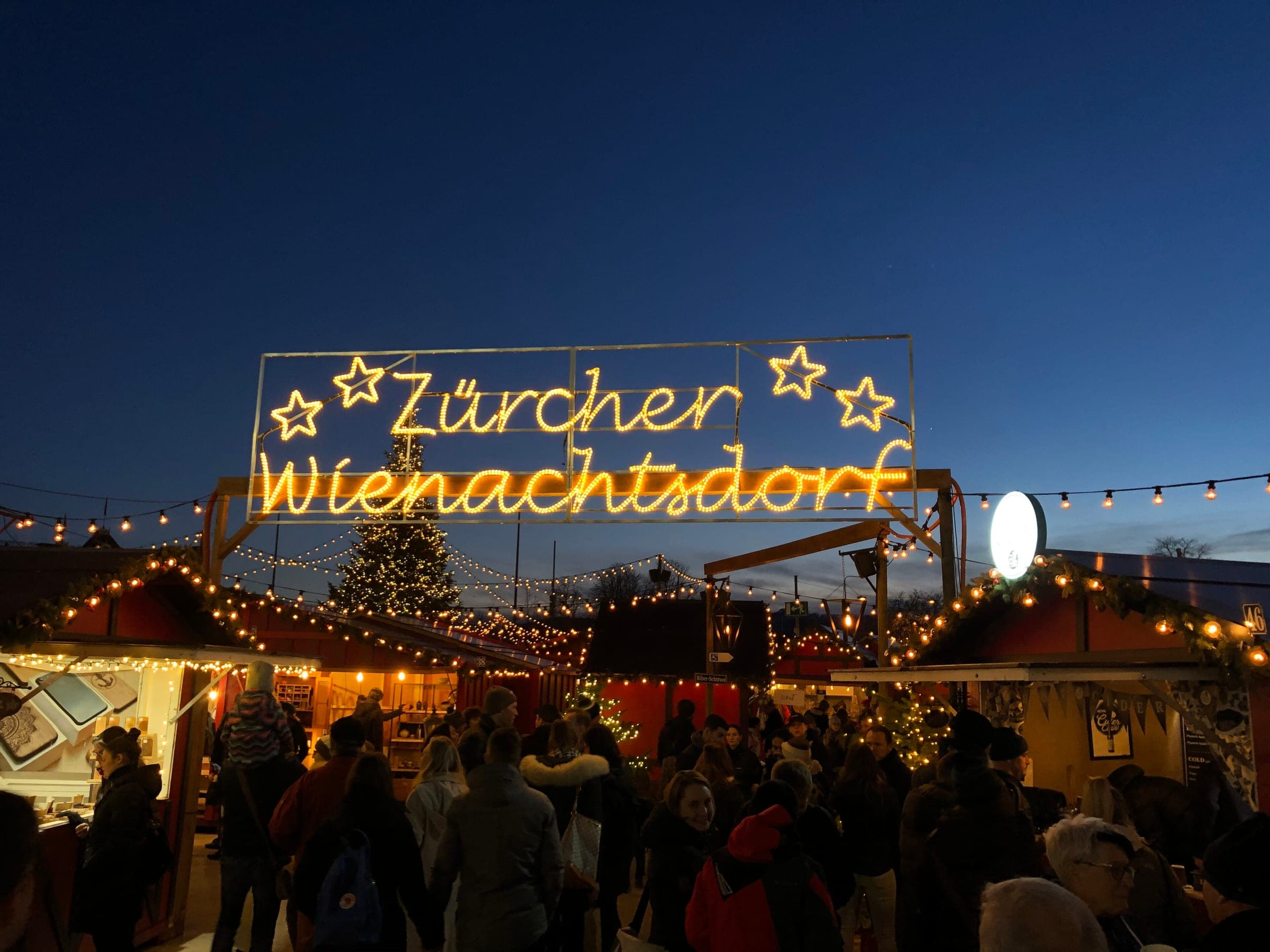 A lit Zürcher Wienachtsdorf sign hangs over the busy stalls on Sechseläutenplatz in Zurich at dusk