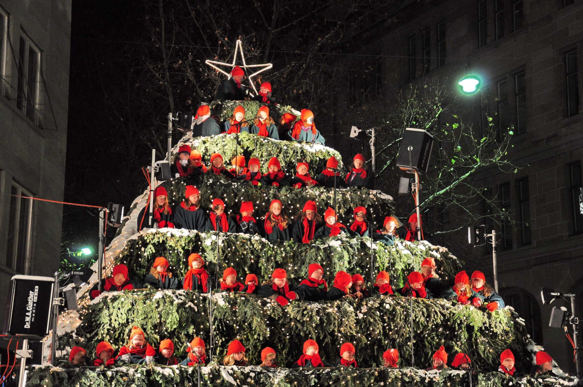 A choir in red hats stands on the tiers of the Singing Christmas Tree in Zurich at night
