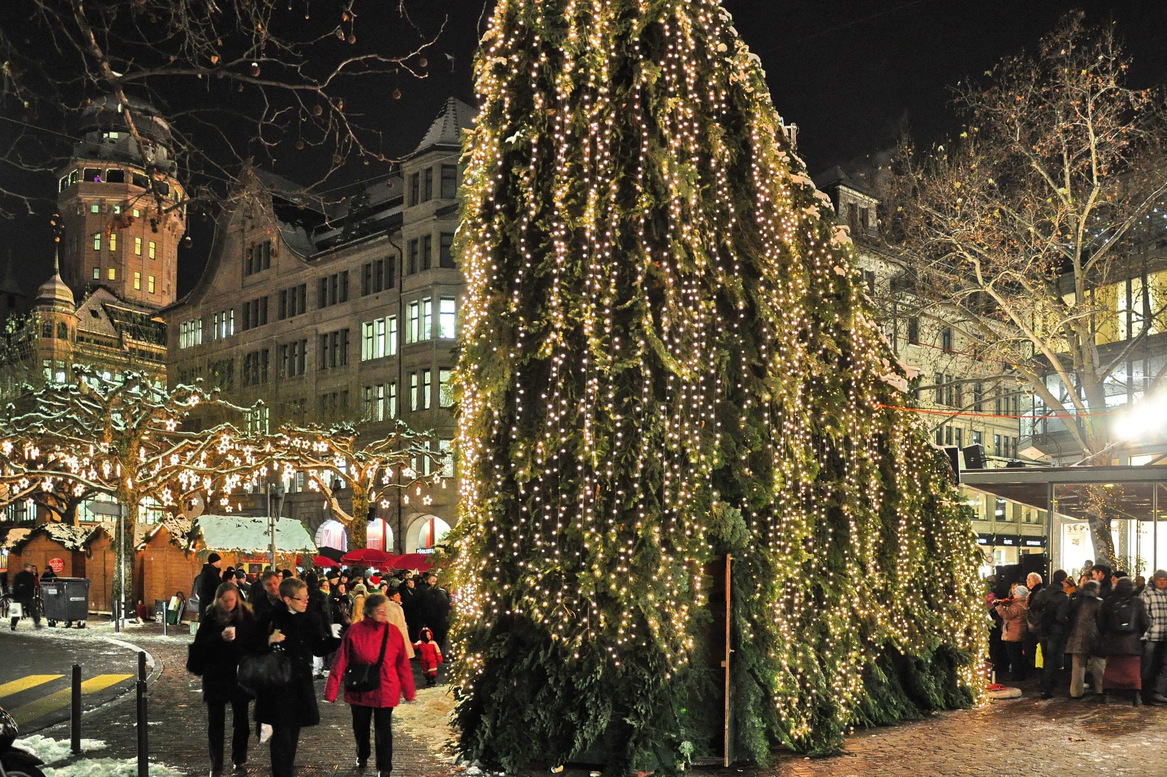 The Singing Christmas Tree glows on Werdmühleplatz in Zurich on a snowy night, with the Urania tower behind