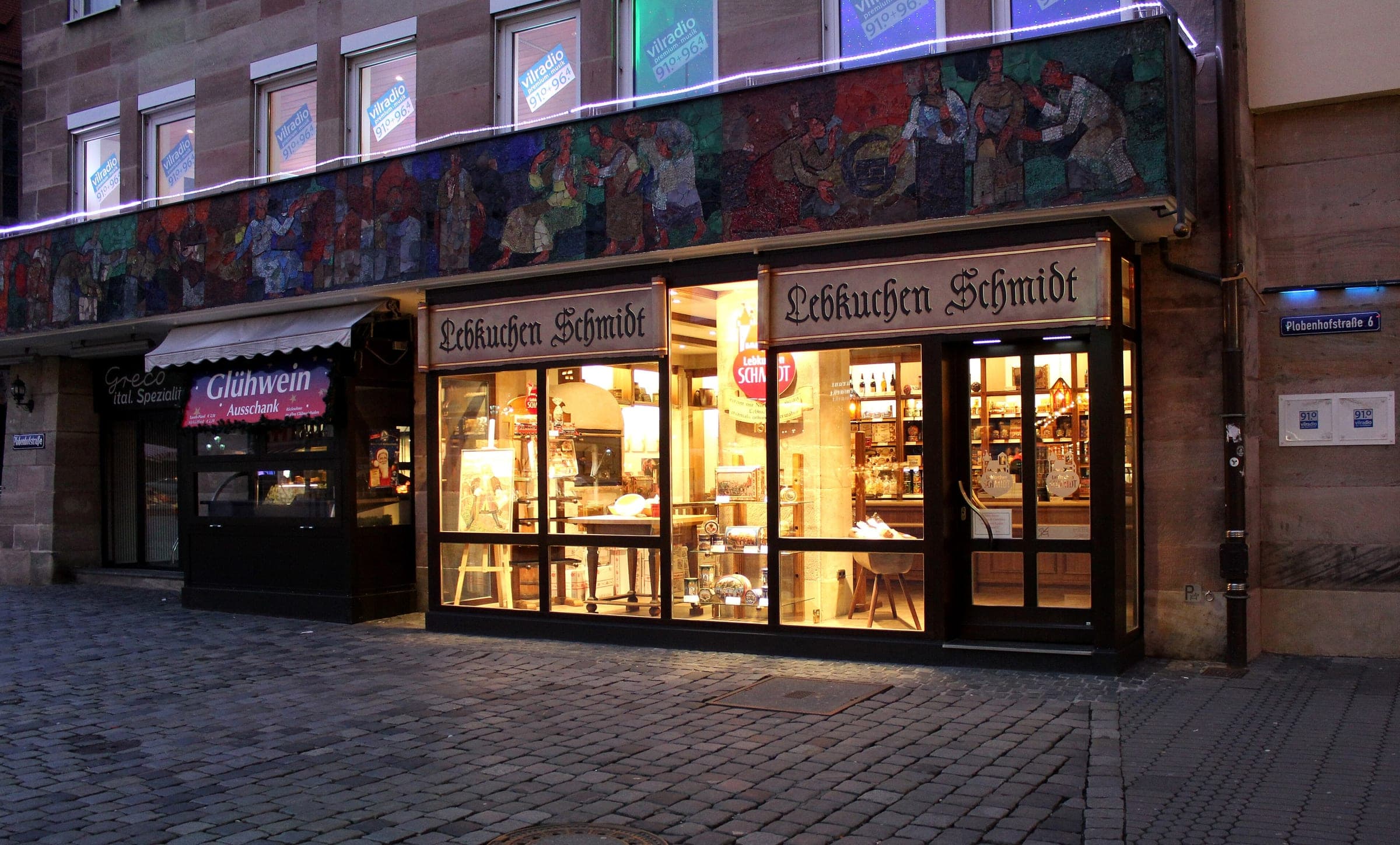The lit shop windows of Lebkuchen-Schmidt glow under a mosaic frieze on Plobenhofstraße in Nuremberg at dusk.