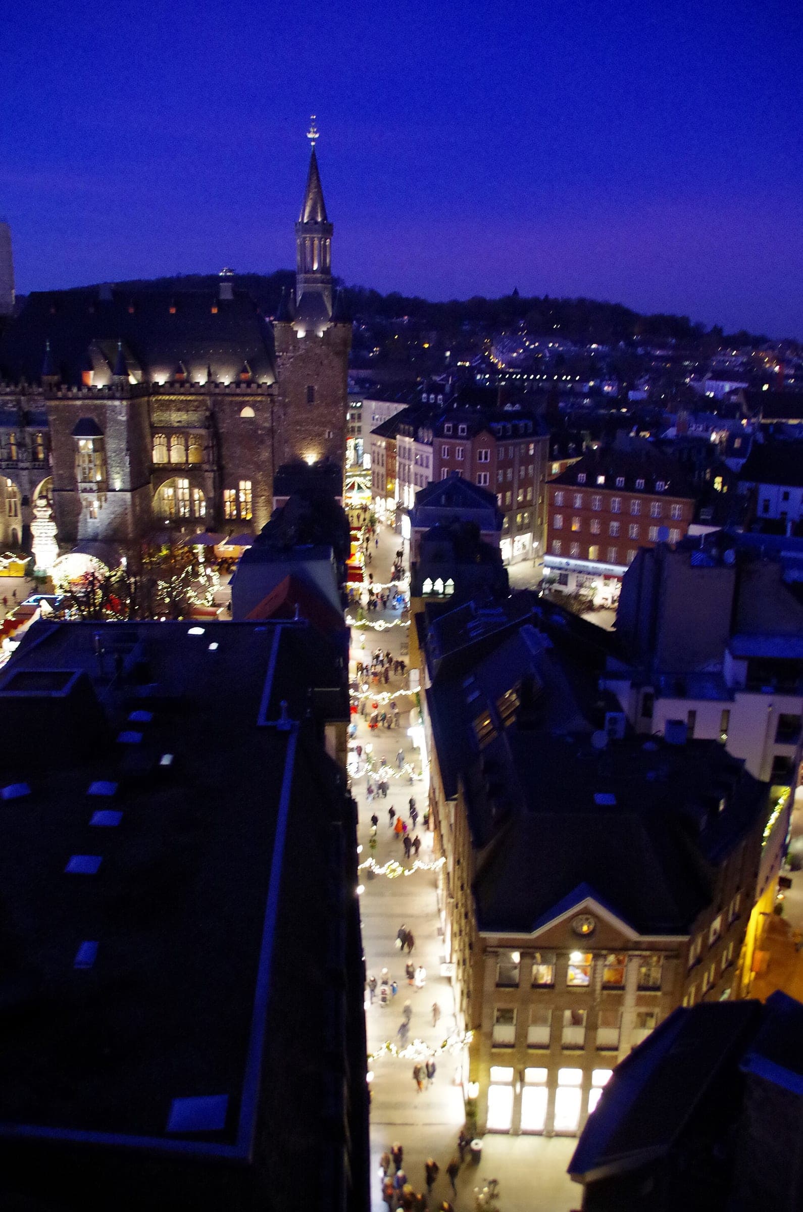 Christmas lights line a crowded street below Aachen town hall at blue hour during the market