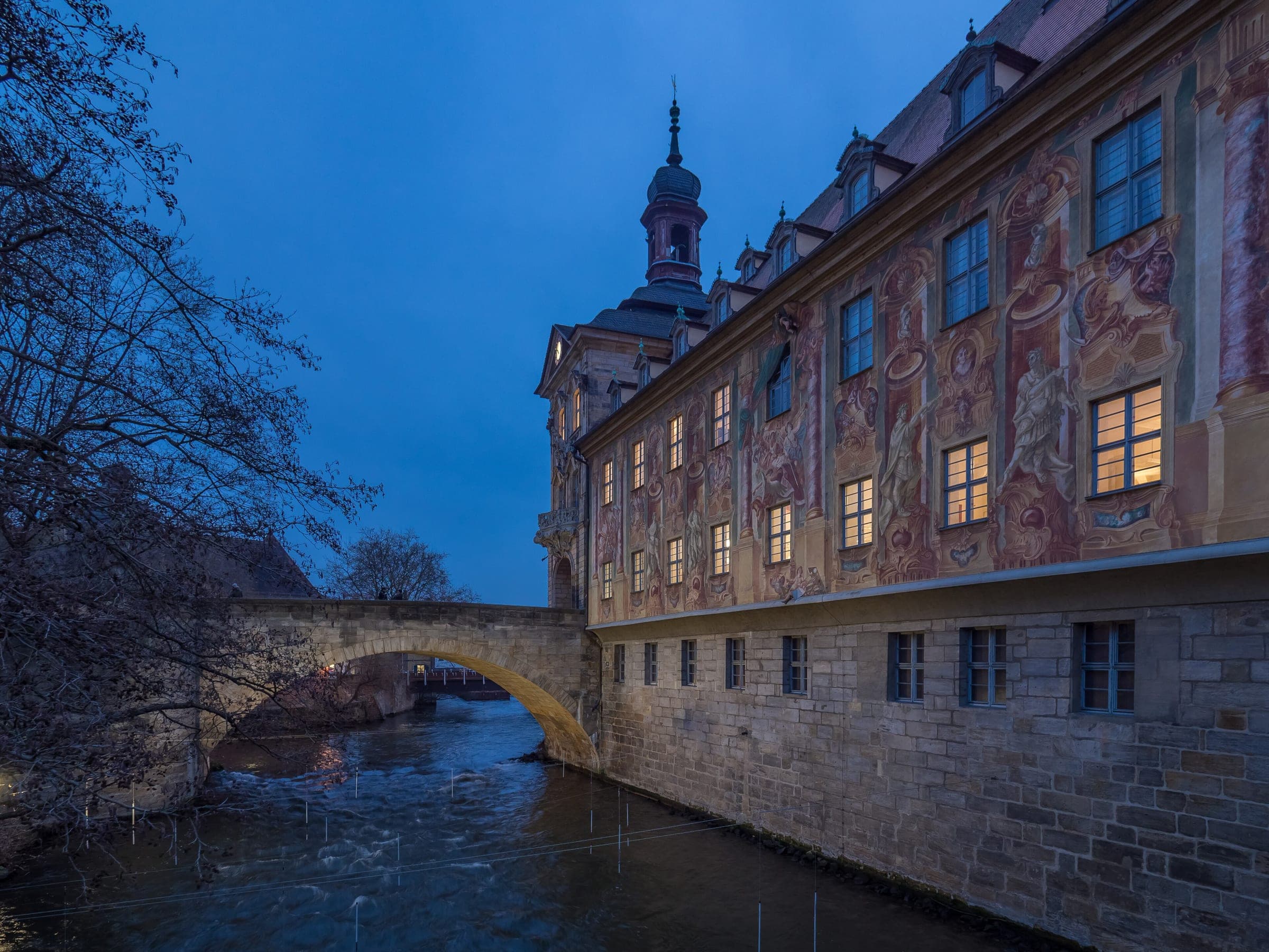 Painted frescoes cover the side of Bamberg's Altes Rathaus above the Regnitz as the windows light up at dusk