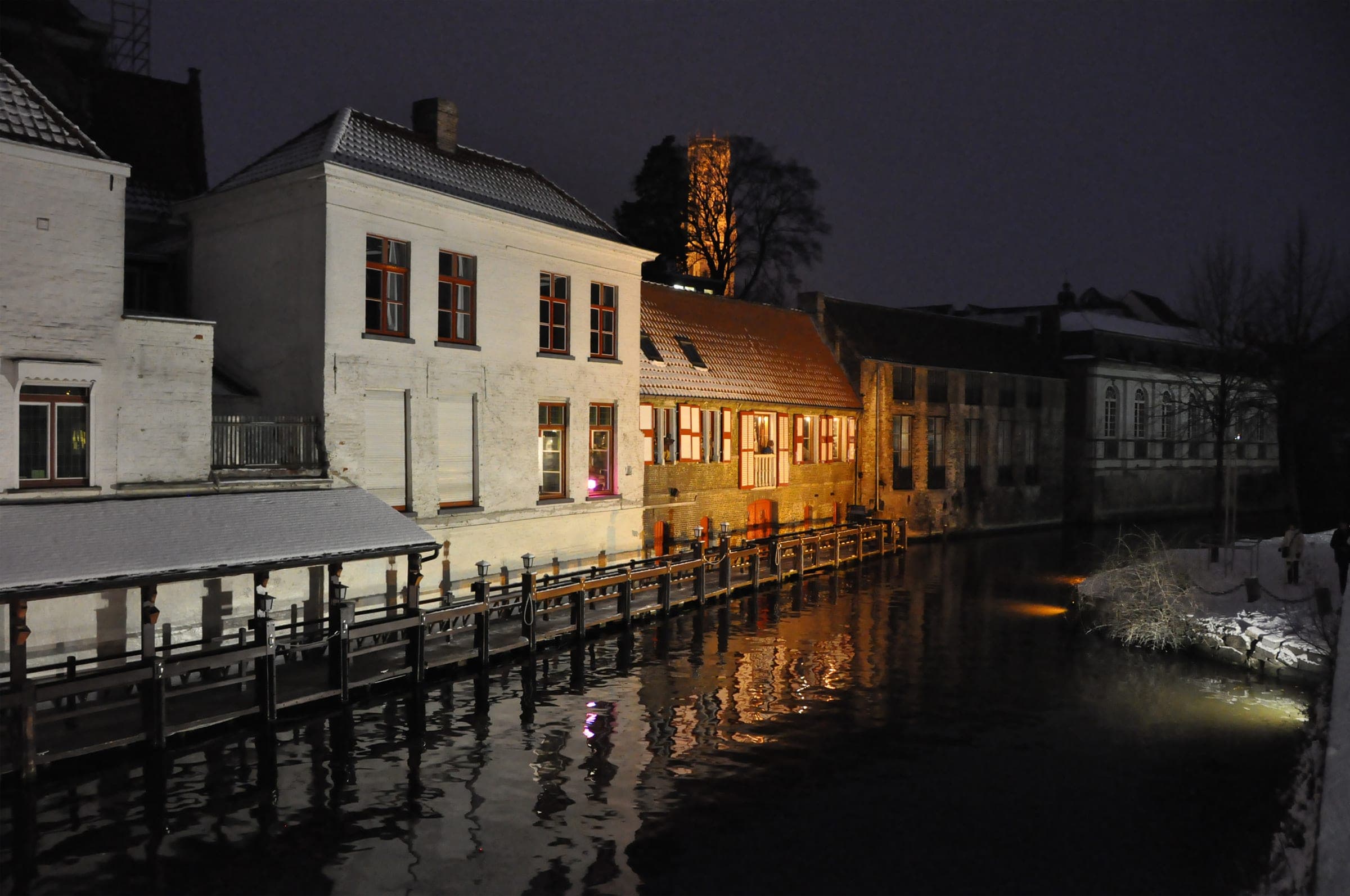 Snow edges the Dijver in Bruges at night while lamps light the old houses along the canal.