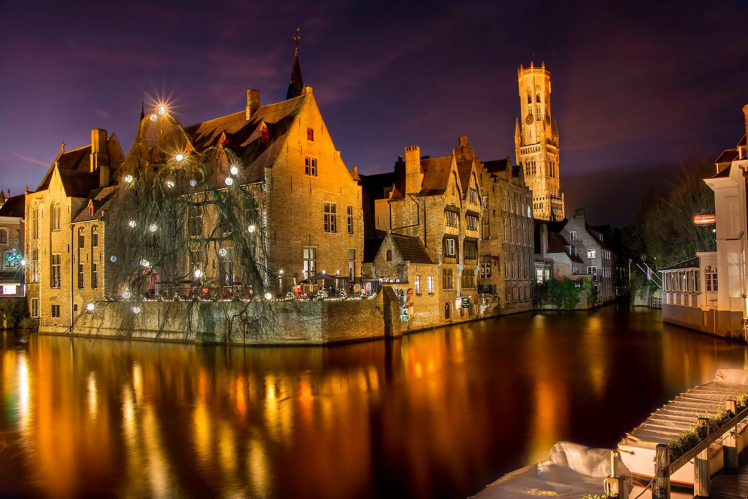 Fairy lights hang in the willow on the Rozenhoedkaai in Bruges as the floodlit Belfry rises over the canal.