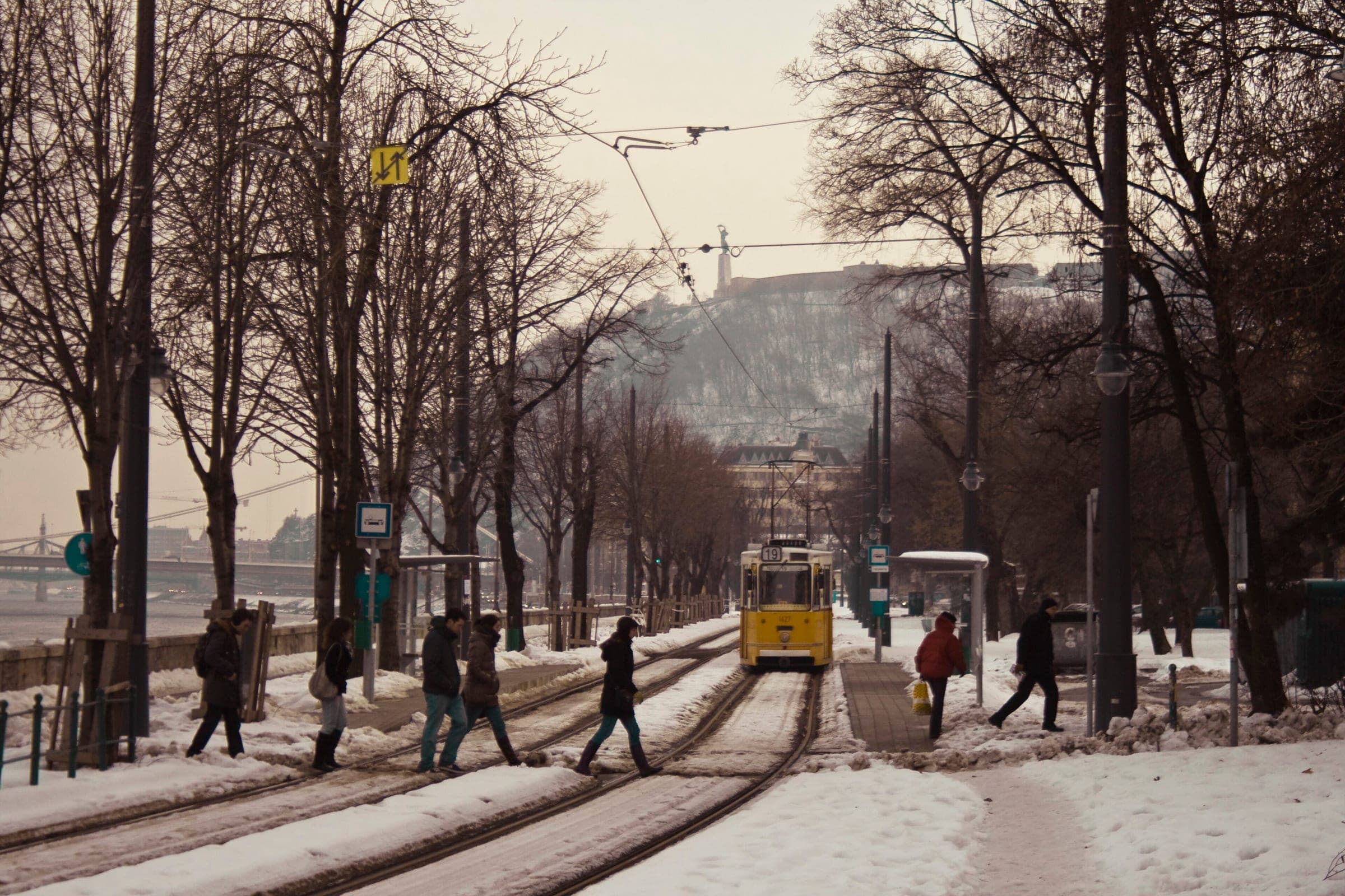 A yellow tram rolls along a snowy tree-lined avenue in Buda with Gellért Hill behind.