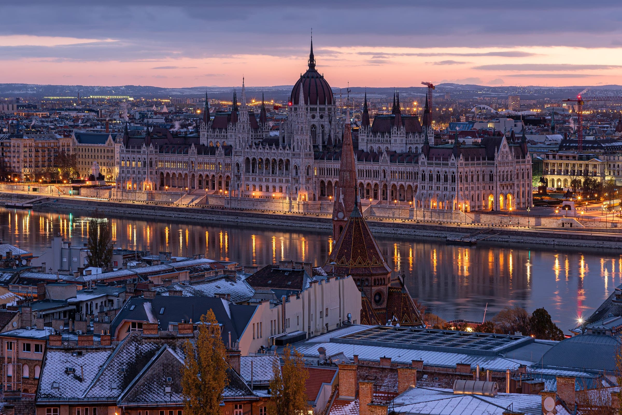 Snowy Buda rooftops lead across the Danube to the lit Parliament in Budapest at dusk.