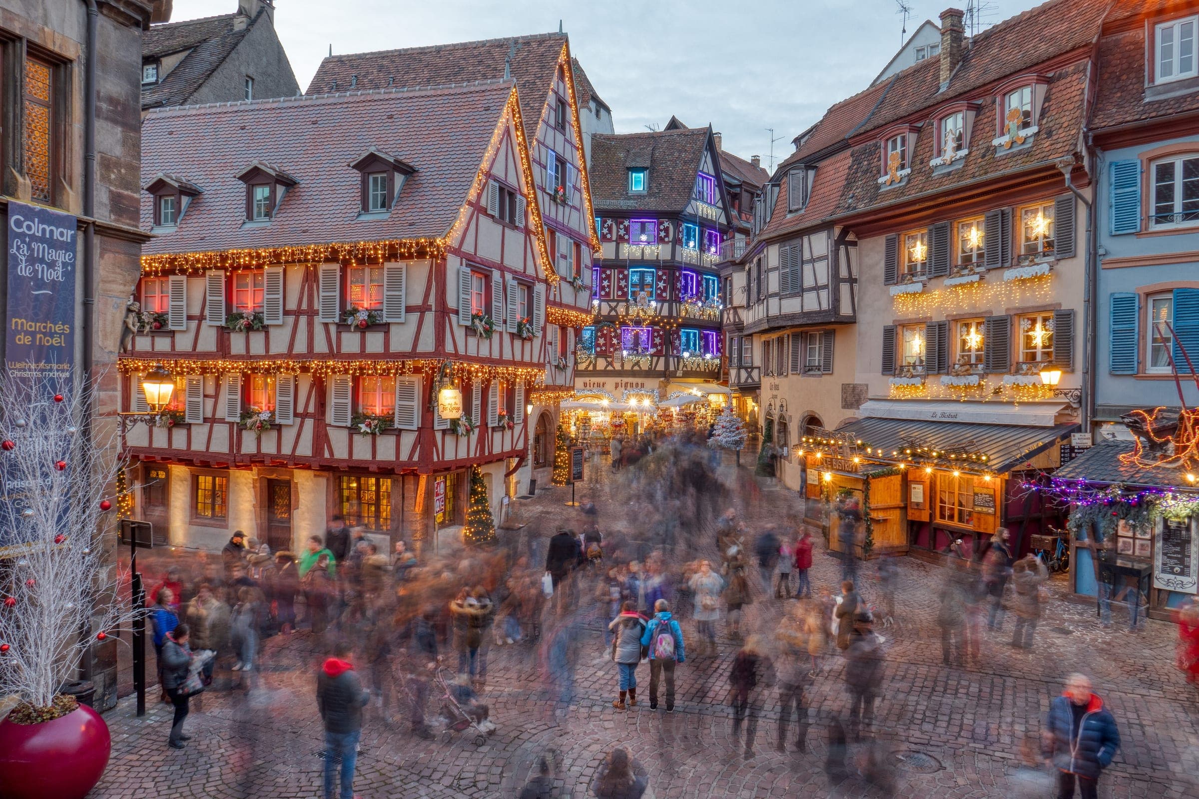 Crowds blur through the old town of Colmar at dusk under half-timbered houses strung with Christmas lights.