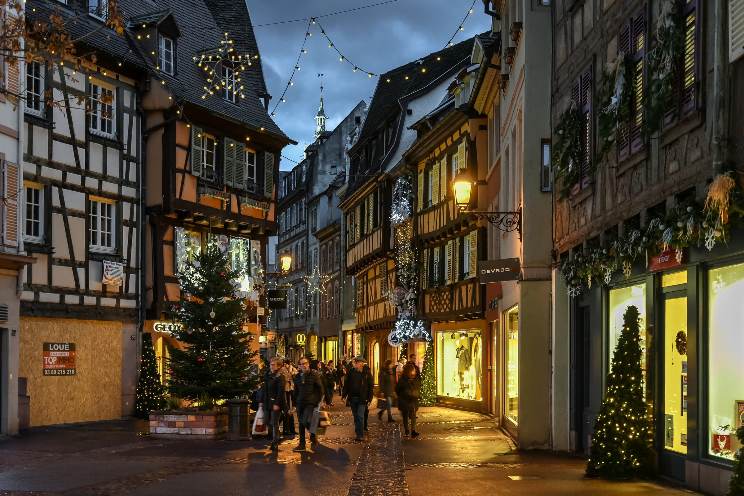 A lit fir stands at a corner of a half-timbered street in Colmar as shoppers pass in the evening.