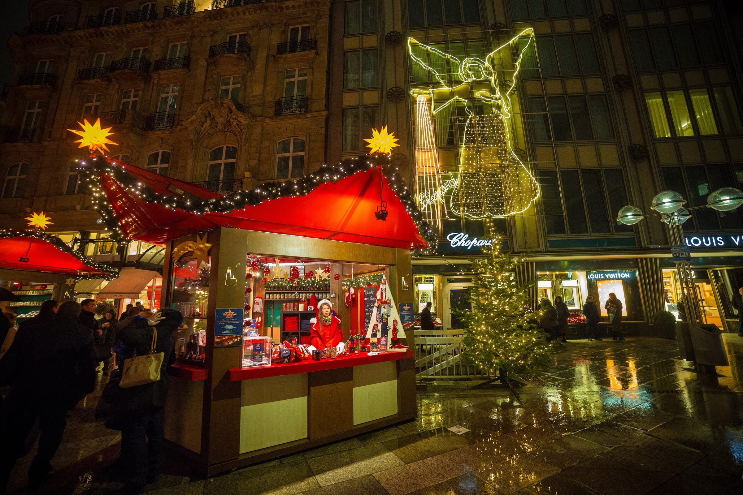 A red-roofed gift stall glows under a giant light angel on a shopping street in central Cologne at night