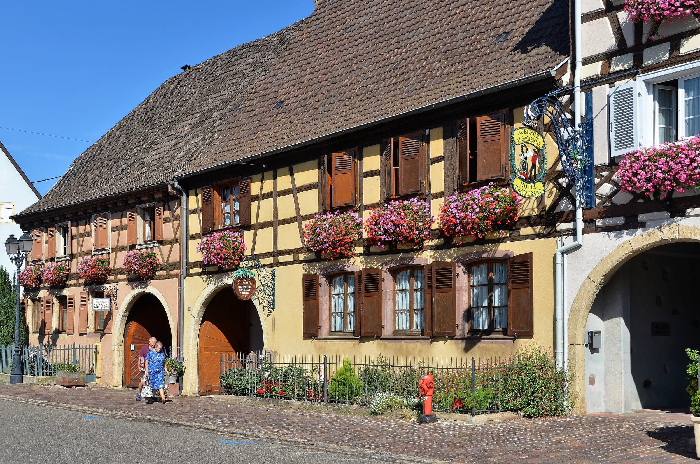 Geraniums spill from the windows of a long half-timbered house beside an arched gateway in Eguisheim.