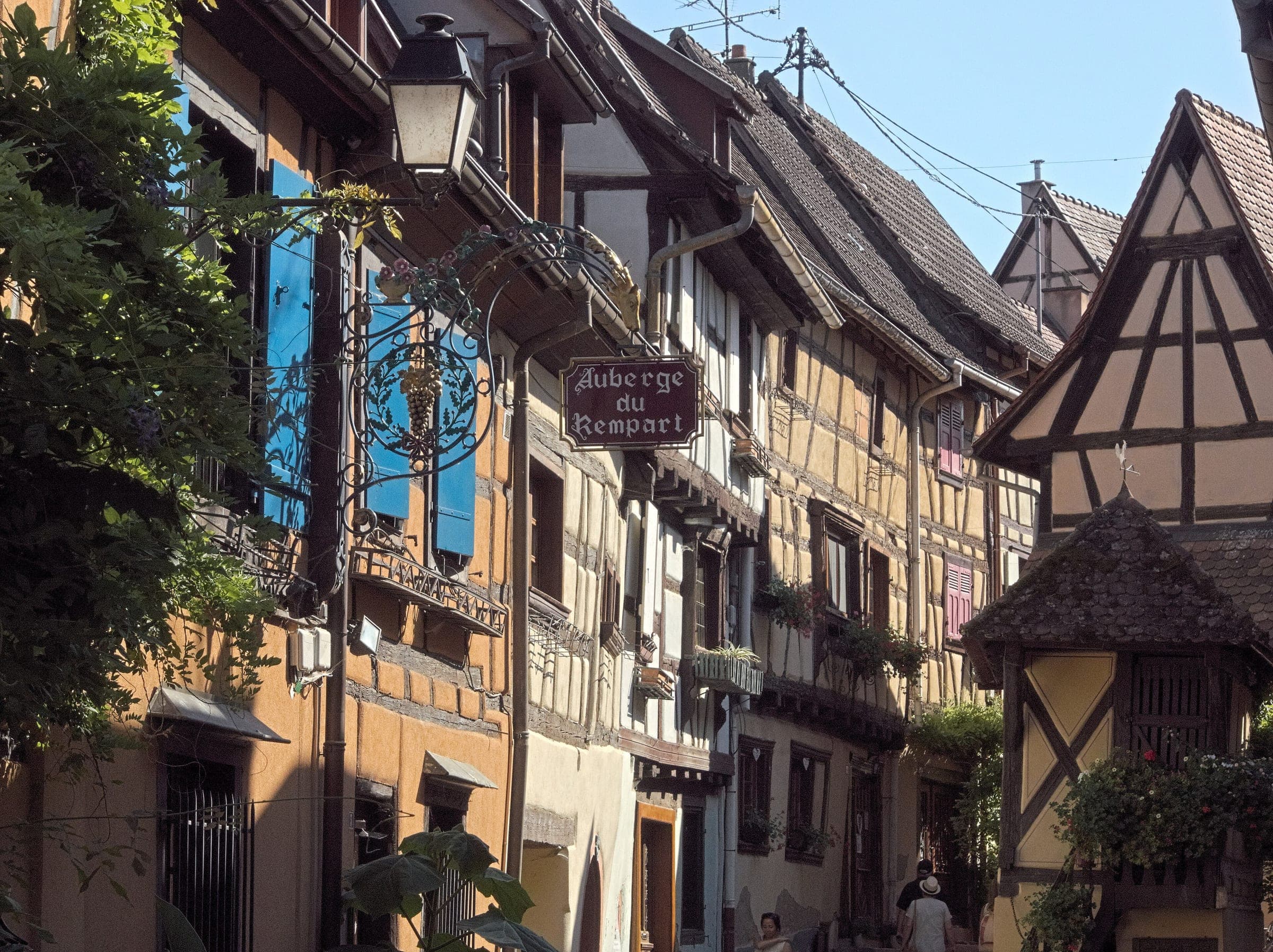Half-timbered houses curve along the rue du Rempart in Eguisheim under a clear sky.