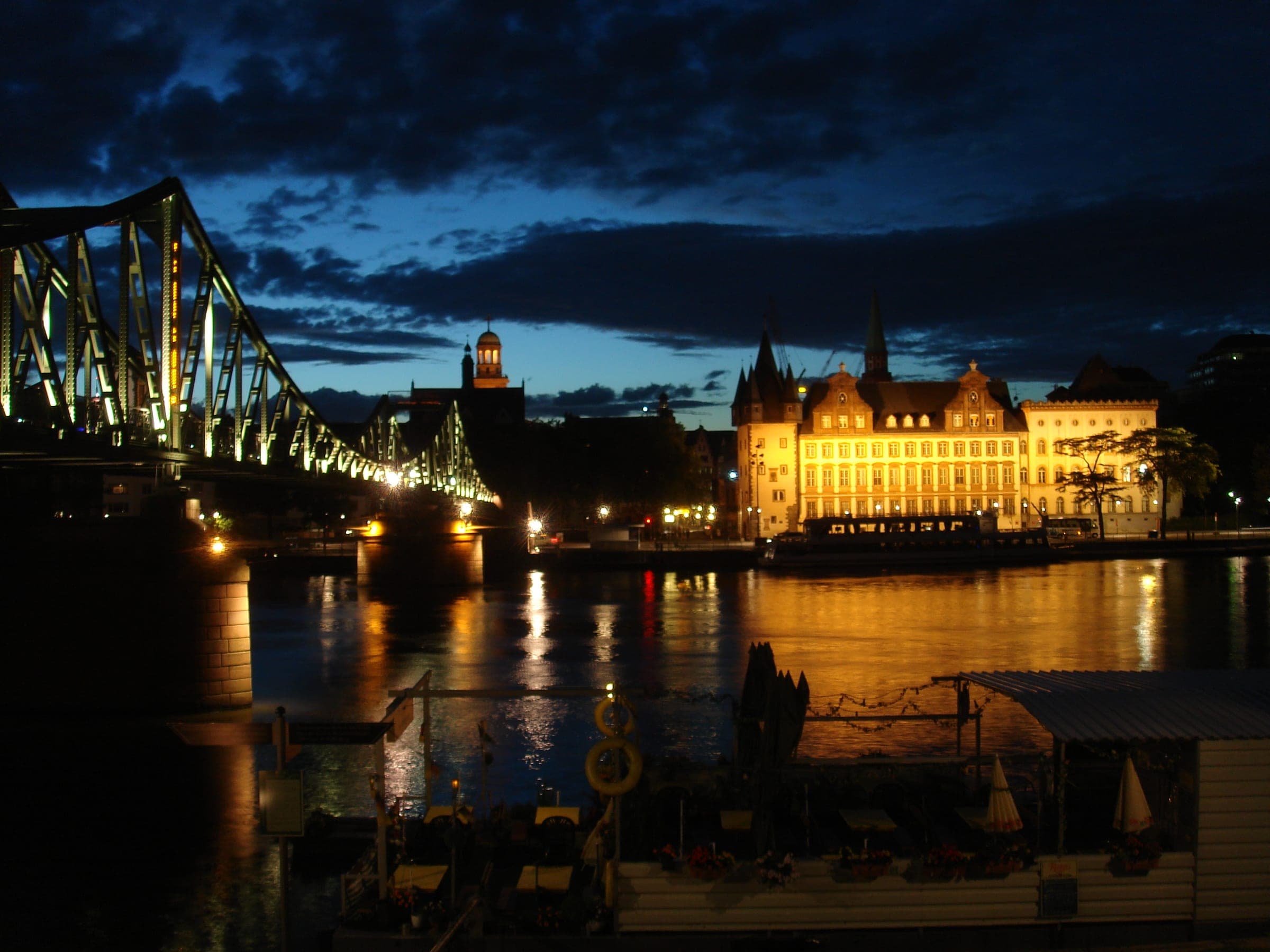 Lights from the Eiserner Steg and the Saalhof shimmer on the Main in Frankfurt under a deep blue evening sky.