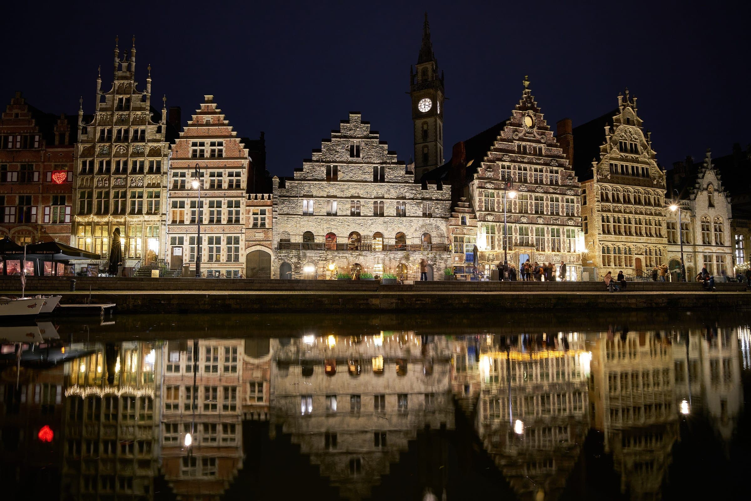 The guild houses of the Graslei in Ghent glow at night and double in the still water of the Leie.