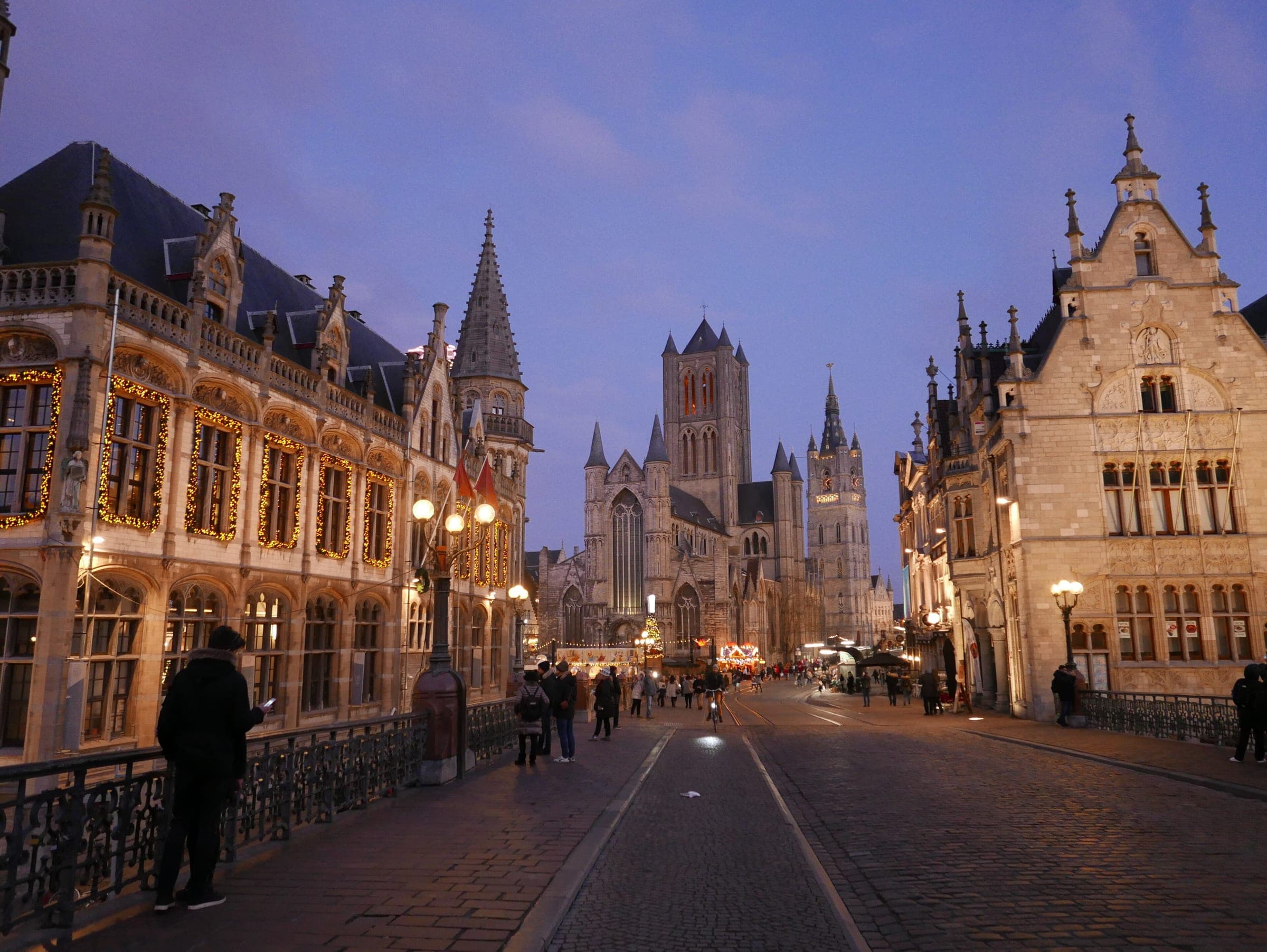 Ghent's three towers rise beyond St Michael's Bridge at blue hour as lights trace the old post office.