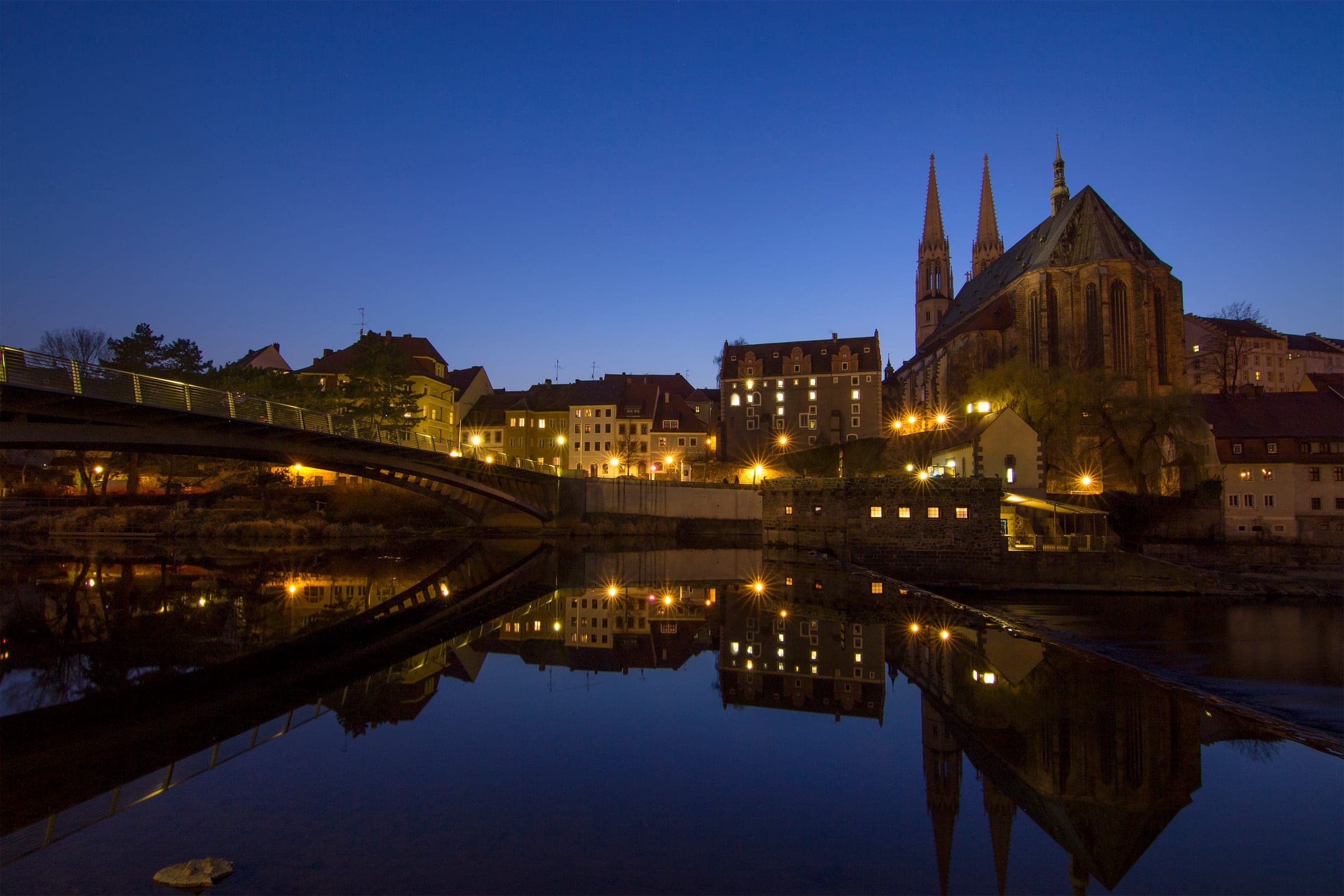 The twin spires of the Peterskirche reflect in the Neisse beside the old town bridge in Görlitz at blue hour.