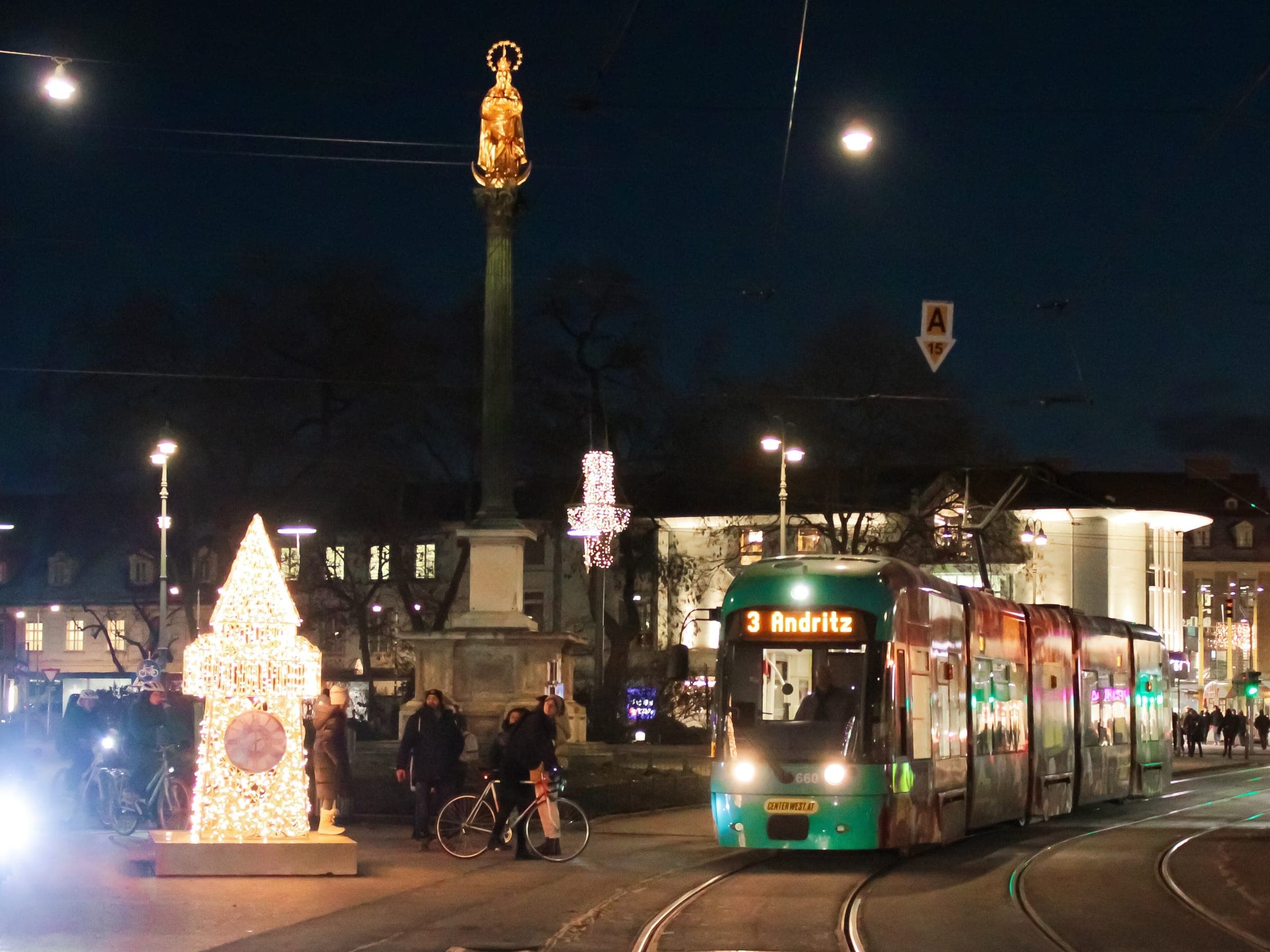 A glowing tree of lights stands by the Marian column at Am Eisernen Tor in Graz as a tram passes at night.