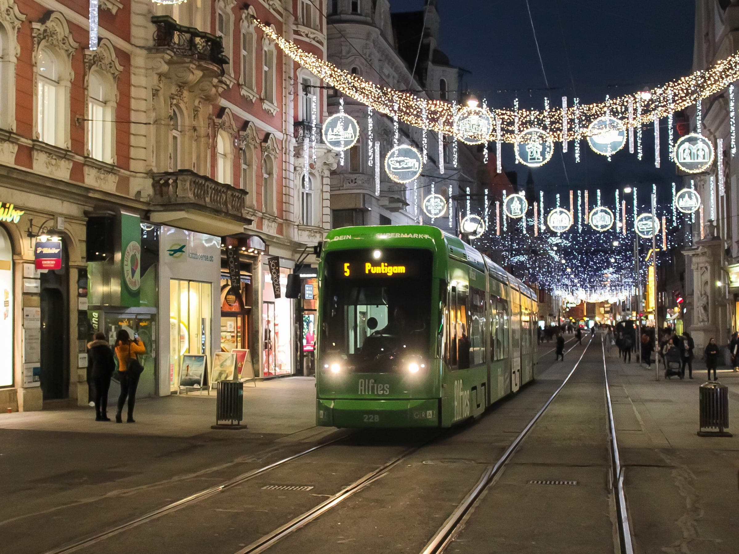 Garlands and hanging baubles light Herrengasse in Graz above a green tram on a December night.