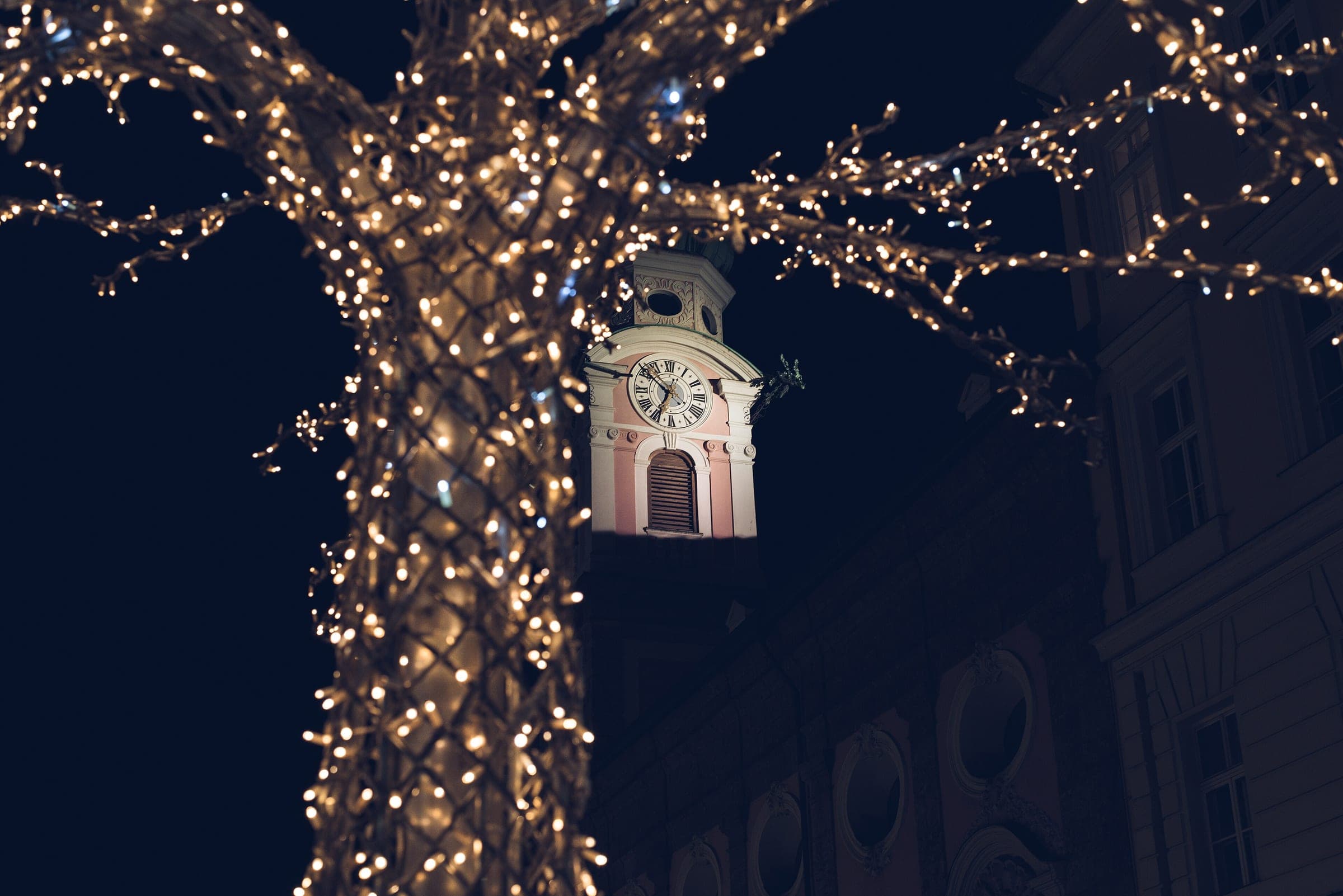 A tree wrapped in fairy lights frames the clock tower of the Spitalskirche on an Innsbruck December night.