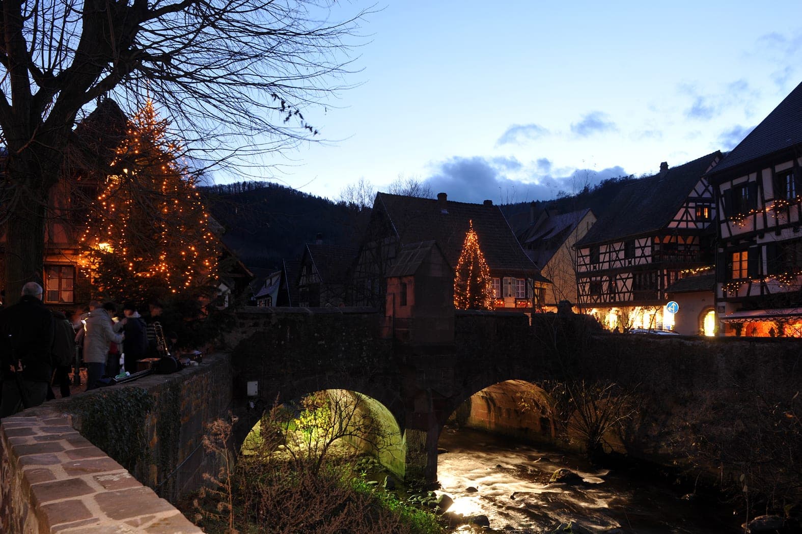 Lit Christmas trees stand by the old stone bridge over the Weiss in Kaysersberg as dusk falls.