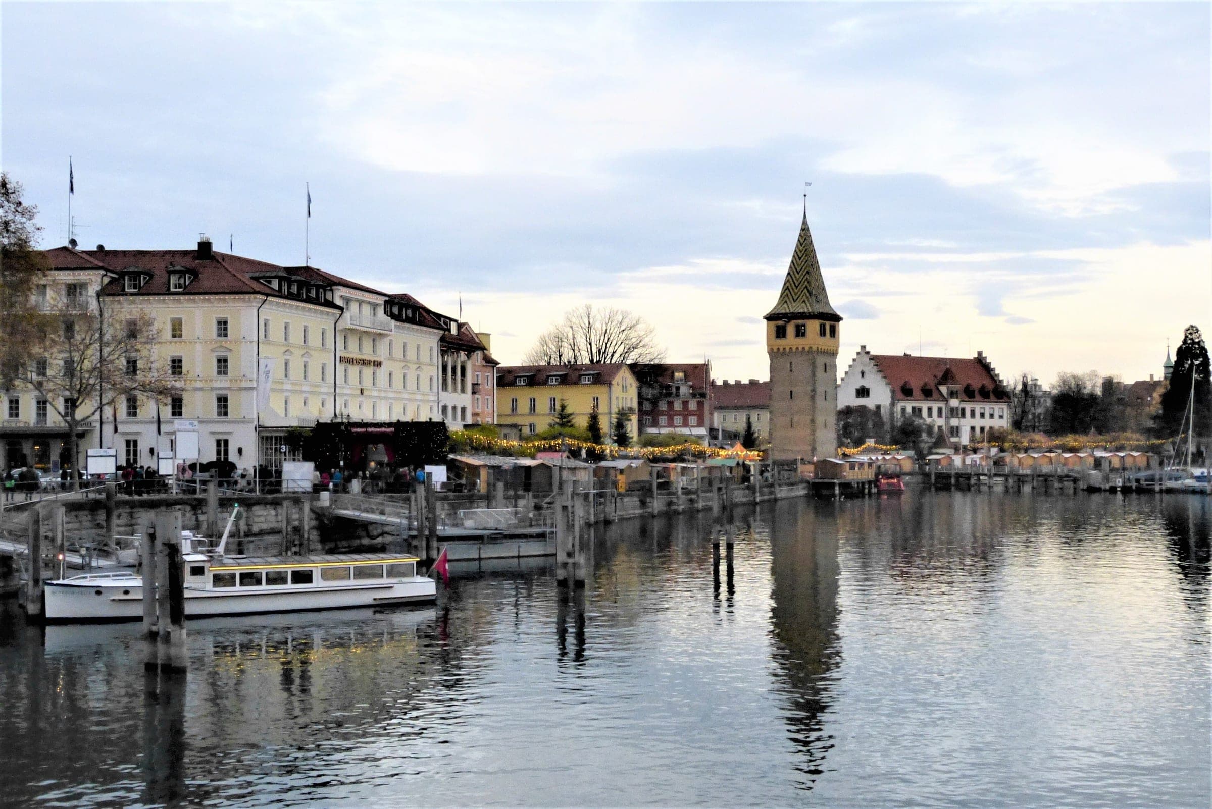 Christmas market stalls line the Lindau harbour quay beside the Mangturm with boats moored in front.